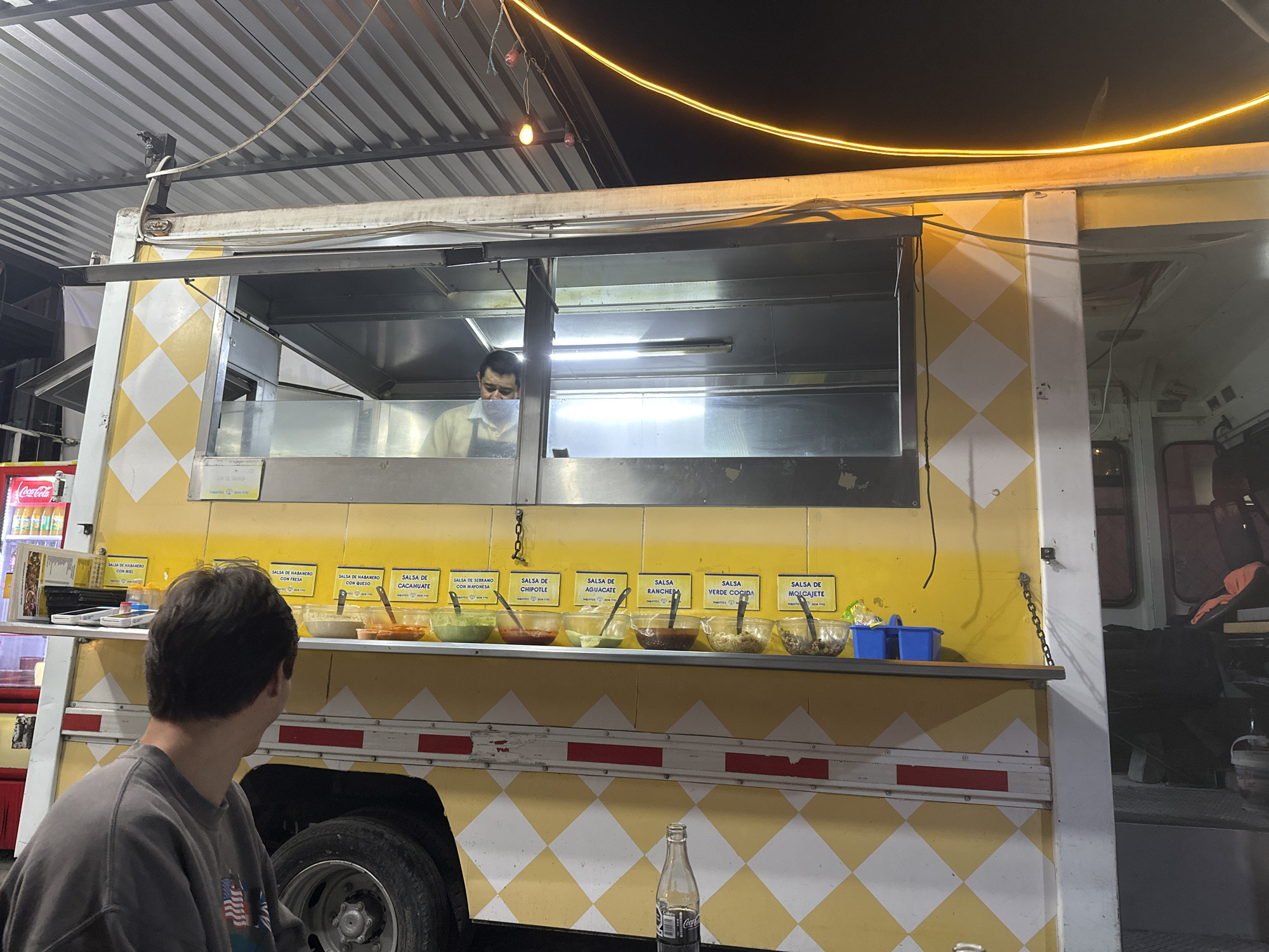 A night scene captures a customer, viewed from behind, seated in front of a vibrant food truck in Puebla, Mexico. The food truck, painted in a distinctive yellow and white diamond pattern, features an open service window where a chef, wearing a face mask, is visible. Above the counter, a prominent display of various salsas in clear glass bowls includes "SALSA DE HABANERO CON MIEL," "SALSA DE HABANERO CON FRESA," "SALSA DE HABANERO CON QUESO," "SALSA DE CACAHUATE," "SALSA DE SERRANO CON MAYONESA," "SALSA DE CHIPOTLE," "SALSA DE AGUACATE," "SALSA RANCHERA," "SALSA VERDE COCIDA," and "SALSA DE MOLCAJETE." The truck also bears signage for "TAQUITOS DON TITO."
The setting is illuminated by internal fluorescent lights within the truck and warm string lights strung overhead, indicating a casual, evening dining experience. A Coca-Cola bottle rests on the counter, and a Coca-Cola refrigerator is partially visible to the left, displaying the brand's logo. A corrugated metal roof extends above the truck, under which the scene unfolds. The customer appears to be observing the scene, possibly awaiting an order from this well-stocked food truck.