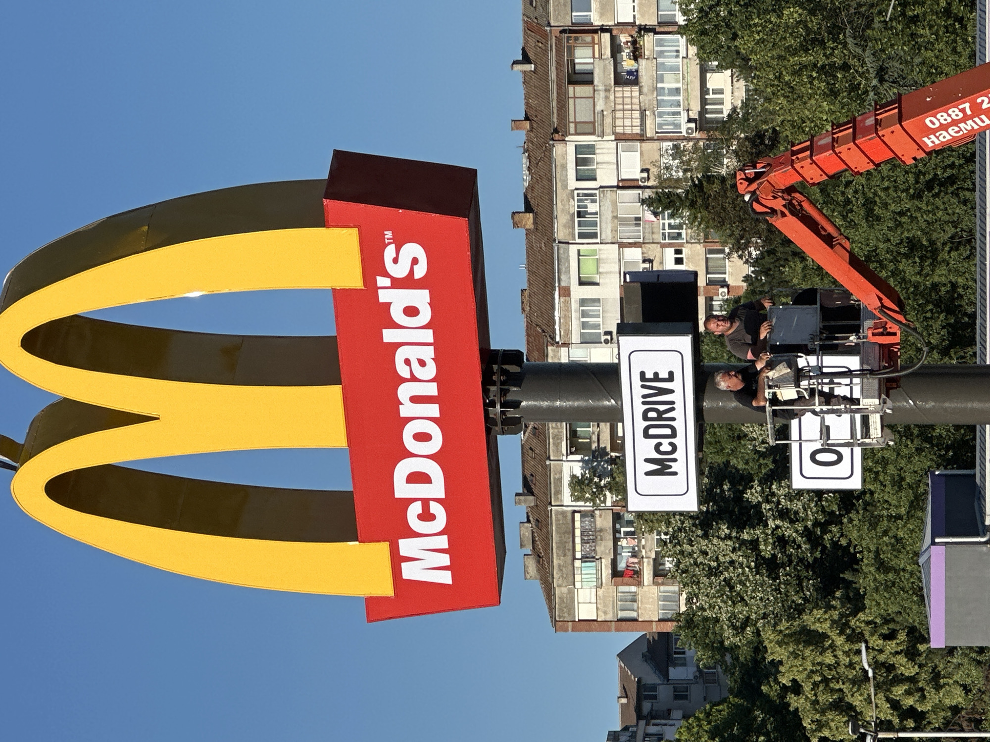 The image shows a large McDonald's sign with the golden arches and red base. Below the sign, there are two white rectangular signs. The upper one says "MCDrive" in black text. The lower one contains a black "O" and then some illegible text. Two men are visible in a cherry picker lift attached to the pole supporting the signs, suggesting maintenance or installation work is underway. In the background, there are apartment buildings and trees. The scene is illuminated by bright sunlight, indicating daytime. The bottom right of the image shows a portion of an orange construction vehicle with Cyrillic text that reads "0887 2..." and "наеми" which translates to "rentals". This specific McDonald's location is on a main road in the city of Veliko Tarnovo, Bulgaria.