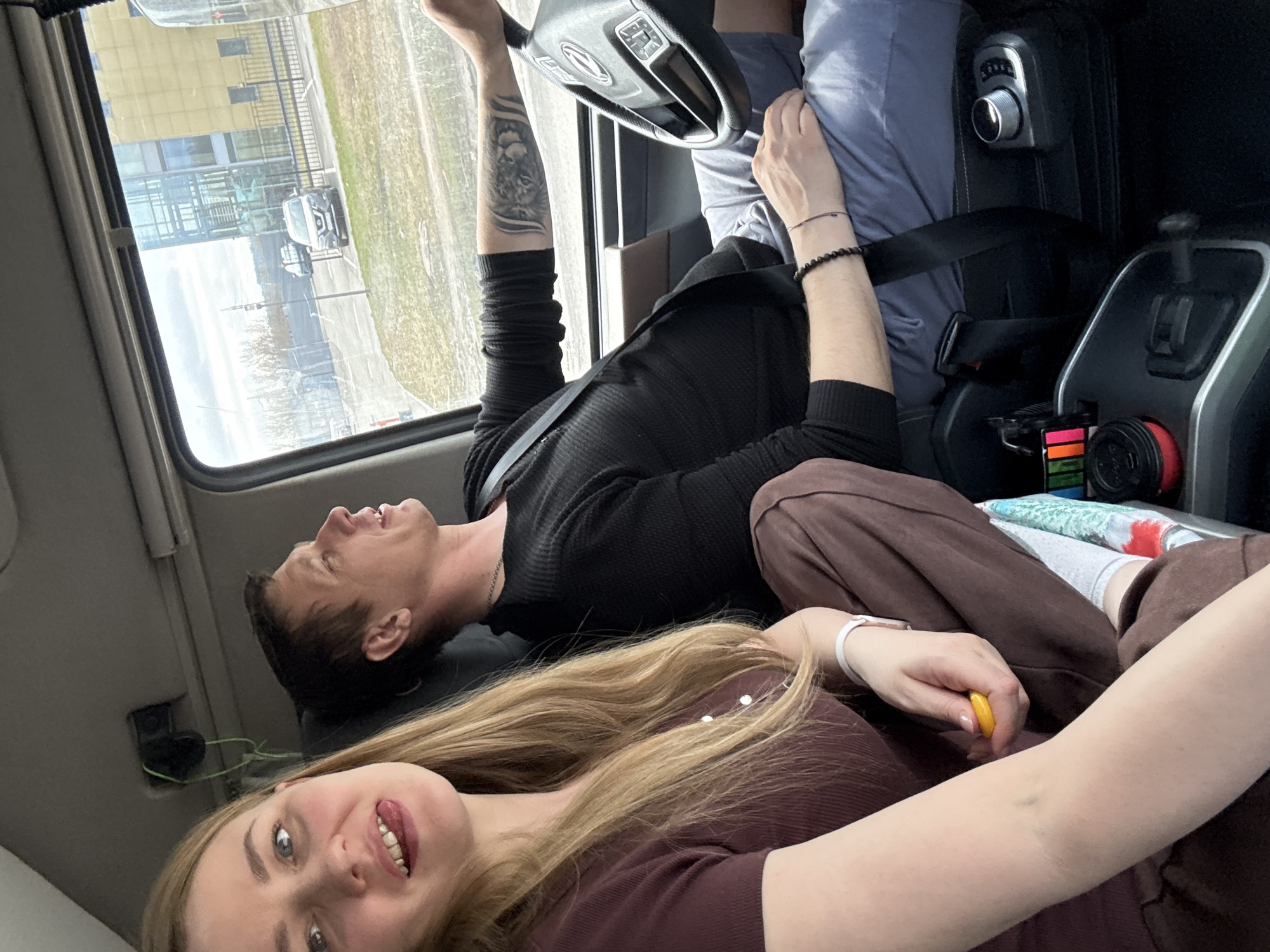 The image appears to be taken inside a vehicle, likely a car or van, with a man and a woman visible. The man is wearing a black shirt and has a tattoo on his left arm. He seems to be lying down, potentially in a reclining seat or across the seat. The woman is in the foreground, smiling and looking at the camera. She has long blonde hair and is wearing a brown shirt. The vehicle is presumably parked or in motion, with a view of buildings and parked cars visible through the window. The scene is likely in Хохряки, Russia, given the prompt's instructions, but there's no specific information in the image to explicitly confirm that. The center console of the vehicle is visible, showing gear shift, a drink cup, and possibly stickers or a small container with various colors. The man is wearing a seatbelt. The woman is wearing an orange ring.