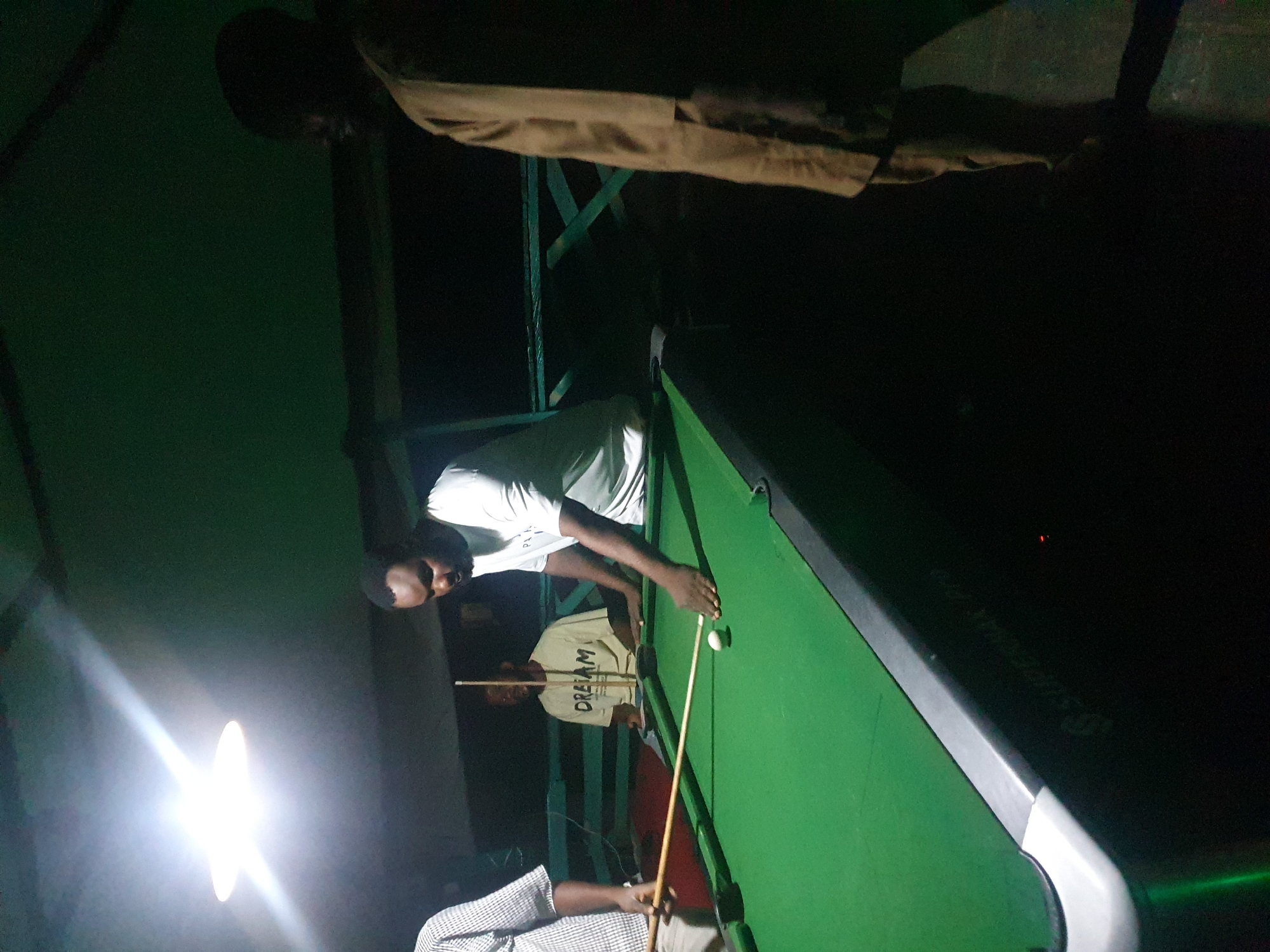 Four men are gathered around a green billiard table in an outdoor, dimly lit setting, likely a bar or recreational area, possibly in Lokoja, Nigeria. The light source, a bright ceiling lamp, illuminates the scene from the upper left. The man in the foreground on the right is wearing a light brown traditional outfit and is partially visible from the waist down, his stance suggesting he is observing the game. To his left, another man in a white t-shirt is actively playing, bent over the table with a cue stick in hand, poised to strike a white billiard ball. A third man, wearing a light-colored t-shirt with "DREAM" printed on it, sits in the background, facing the table. The fourth man, on the far left and mostly out of frame, is partially visible and appears to be holding a cue stick. The billiard table has a grey metal edge and a dark green felt surface. The background consists of dark, indistinct structures and fencing.