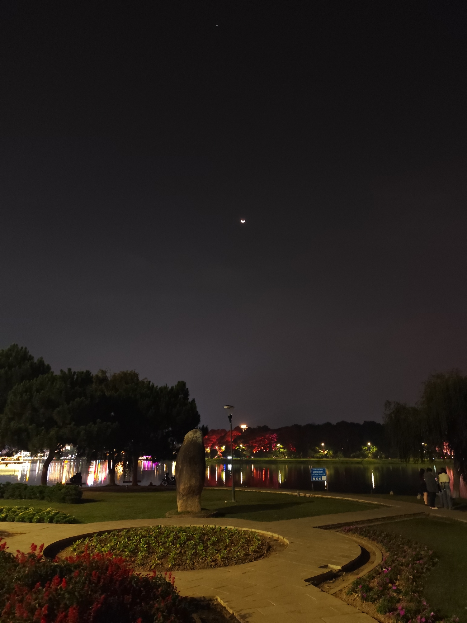 The image captures a serene night scene at a park or lakeside area in Da Lat, Vietnam. The sky is dark, featuring a distinct crescent moon in the upper center and a single bright star or planet in the top left.
In the foreground, a winding paved pathway, possibly made of light-colored stone or concrete, curves through manicured garden beds adorned with vibrant red flowers and other green plants. Grassy areas are also visible on either side of the path. A large, dark, upright stone monument or sculpture stands prominently to the right of the path, near a lamppost.
The midground features a calm body of water, likely a lake or pond, reflecting the colorful artificial lights from the opposite bank. These lights, appearing as streaks of red, purple, white, and yellow, illuminate the trees and structures lining the far shore, creating a striking visual effect on the water's surface. On the near side of the lake, on the left, several large trees stand, one of which shelters a bench where two people are seated. To the right, near the water's edge, two or three individuals are standing, observing the scene. A blue sign by the water, partially obscured but legible from closer inspection, reads "CẢNH BÁO NGUY HIỂM" (Warning Danger) and "CẤM CÂU CÁ" (No Fishing), indicating regulations for the area.
The background consists of a dark treeline silhouetted against the dim night sky. The overall atmosphere is peaceful and contemplative, with the interplay of natural celestial bodies and artificial lights providing a picturesque evening landscape.