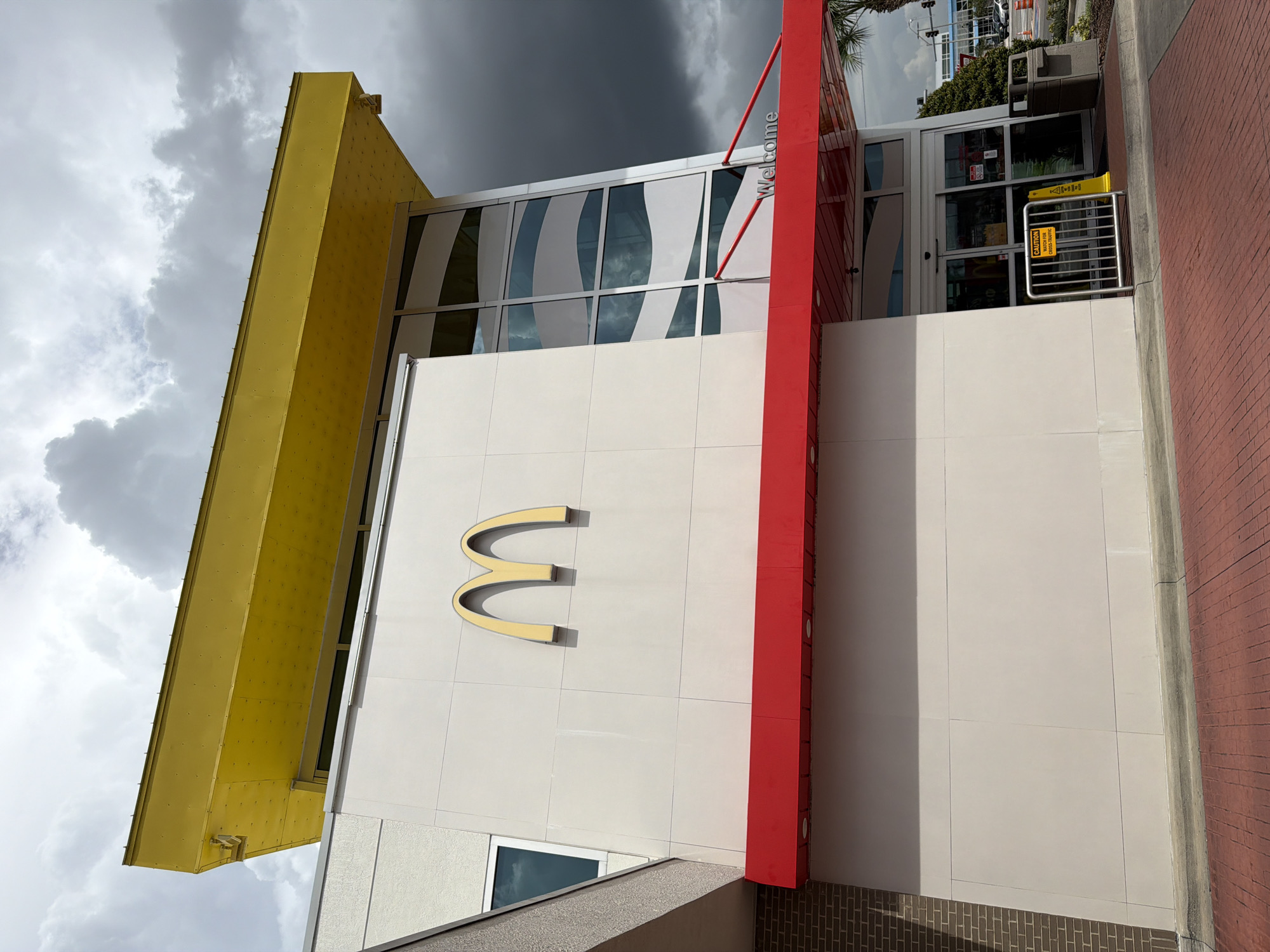 A McDonald's restaurant, located in Tangelo Park, United States of America, is captured from a low angle, emphasizing its modern architecture. The building features a prominent bright yellow overhang, a light-colored facade with the iconic golden arches logo, and a red awning extending over the entrance. Large glass windows with white wavy patterns are visible on the right side of the building. The entrance area has glass doors, a yellow caution sign, and a silver metal barrier with a sign indicating "CAUTION WATCH FOR CROSS-TRAFFIC." A trash receptacle is located to the right of the entrance. The ground in front of the building is paved with red bricks, and a sidewalk separates it from a street. The sky above is filled with dark, heavy clouds, suggesting overcast weather. A palm tree is partially visible to the right of the entrance.