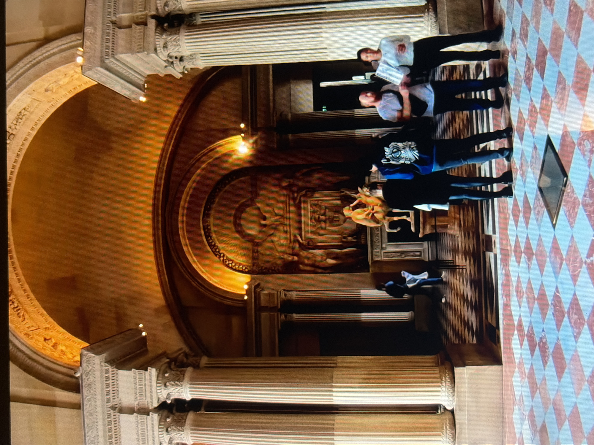 The image depicts the interior of a grand building, likely a museum or gallery, with classical architecture. The scene features a large, arched alcove at the rear, containing a relief sculpture and two statues. In front of this alcove is a bronze sculpture of a figure playing a lyre, positioned on a pedestal. The floor is tiled in a checkerboard pattern of red and white squares.
Several individuals are present in the scene. In the foreground, four people stand facing away from the viewer, observing the artwork. One person in the center wears a dark hoodie with a white graphic on the back. To their right, a man in a white t-shirt and dark pants is visible, with a black fanny pack around his waist. A woman in a white top and dark pants stands next to him. Another person in dark clothing is partially obscured to the left.
Further into the background, a person in dark clothing is seen walking away from the camera. The architecture is characterized by large, fluted columns with ornate capitals, and a decorative archway above the main alcove, illuminated by warm lighting. The overall impression is one of an art exhibition space with visitors admiring historical artifacts. The image is a digital photograph with a slightly grainy texture, suggesting it might be a screenshot or a lower-resolution capture.