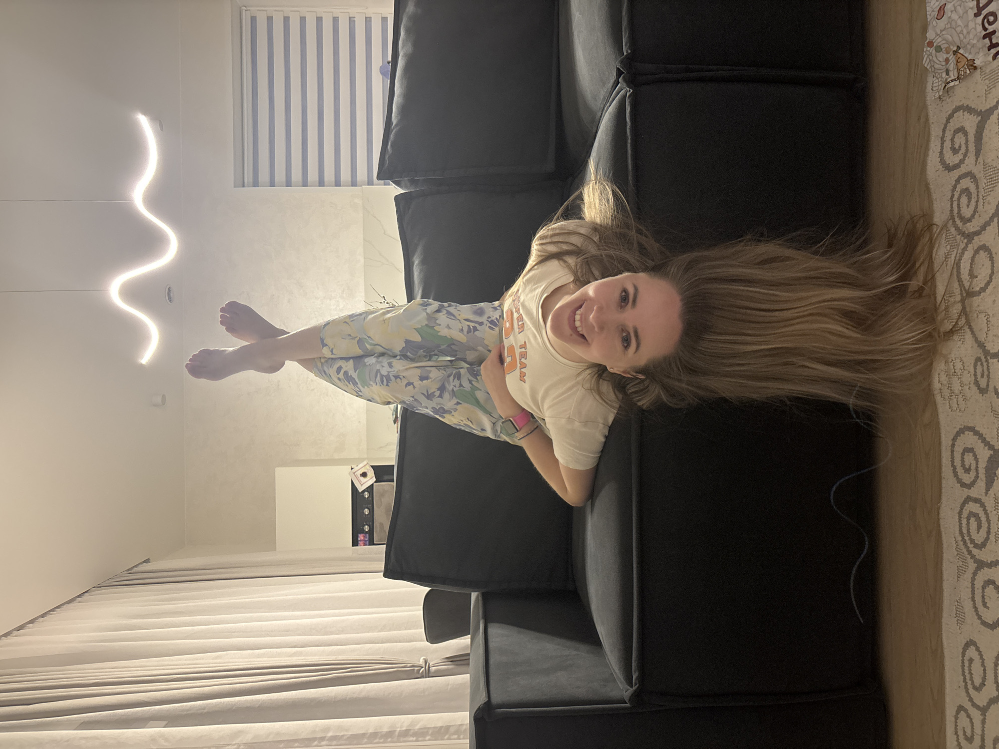 A fair-skinned woman with long blonde hair is reclining on a dark gray couch in a brightly lit interior. She is wearing a light-colored t-shirt and patterned pants. The room includes a modern, wavy light fixture, white blinds, and a rug with a patterned design. A microwave oven is visible in the background. The location is suggested to be Chaikovsky, Russia.