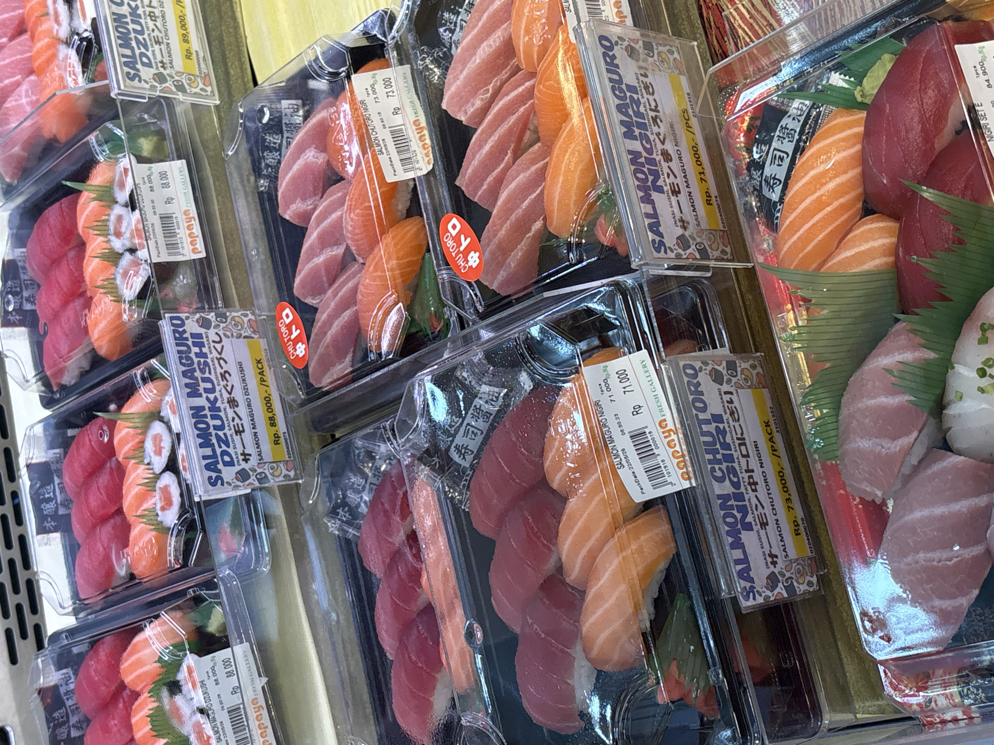 The image shows a display of pre-packaged sushi in clear plastic containers, likely in a supermarket or deli setting. The sushi types include salmon, tuna, and various combinations presented as "Salmon Maguro Dzukushi" and "Salmon Chutoro Nigiri." Prices are indicated in Indonesian Rupiah (Rp), ranging from Rp 71,000 to Rp 89,000. The packaging has branding from "Papaya Fresh Gallery," suggesting a specific store or chain. Text in Japanese is present. The image was likely taken in Medang, Indonesia, as the price tags indicate Indonesian currency.