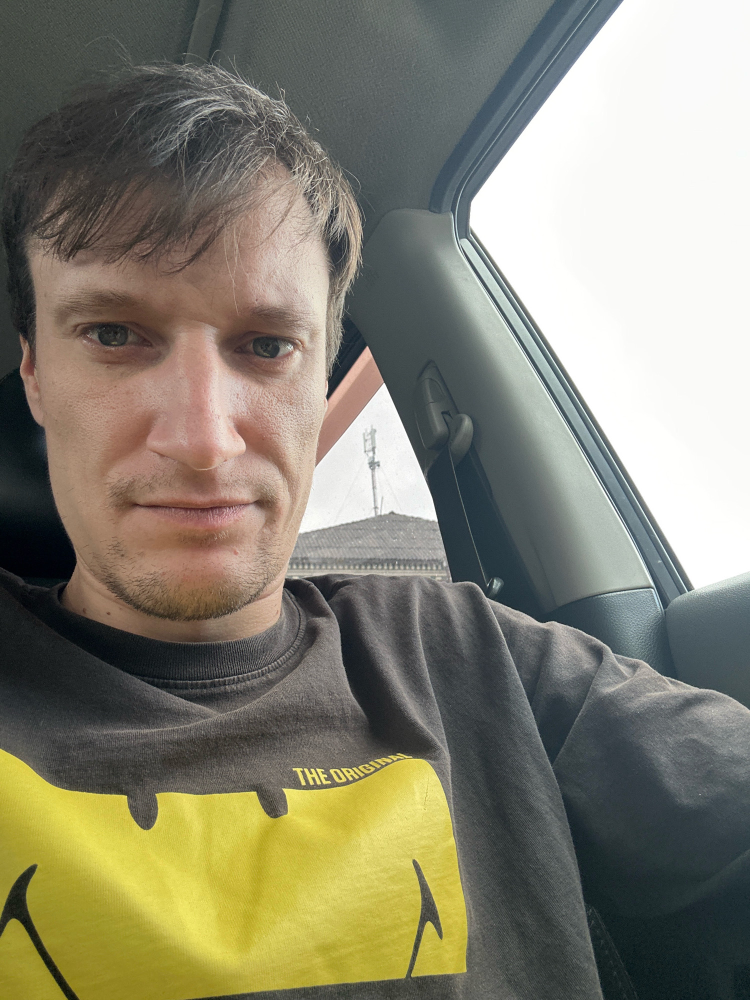A man with fair skin, brown hair, and light eyes is sitting in the driver's seat of a car. He is wearing a dark gray long-sleeved shirt with a large yellow design on the front, possibly a logo or graphic. The car interior is visible, including the dashboard, a seatbelt, and a window. Outside the car, a cloudy sky and a distant building with a roof and what appears to be a cell tower are visible. The man is looking at the camera and taking a selfie. The image does not contain information to determine the precise location within Shatava, Ukraine.