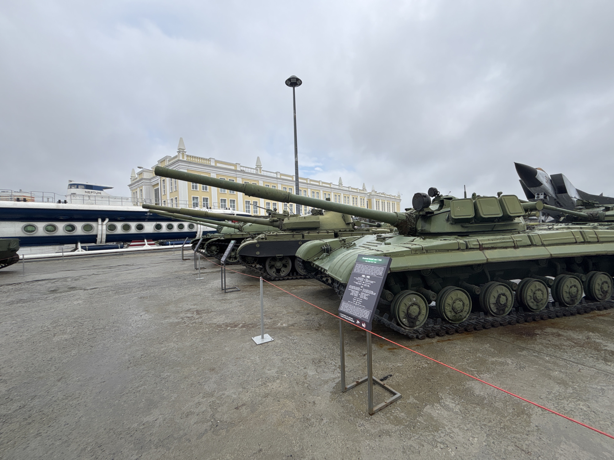 The image displays a military museum exhibition in Verkhnyaya Pyshma, Russia. In the foreground, a row of olive-drab military tanks is parked on a wet, paved surface. The tanks are positioned diagonally, with their tracks and road wheels clearly visible. A red cord runs horizontally in front of the tanks, acting as a barrier. Informational signs are placed in front of some of the tanks, one of which is in focus and displays text. In the background, a white and yellow building with multiple windows stands under a cloudy sky. To the left of the building, a large white ship with round windows, identified by the name "NEPTUN" on its side, is partially visible. A tall lamppost is also present in the mid-ground. The overall scene is an outdoor exhibition of military hardware.