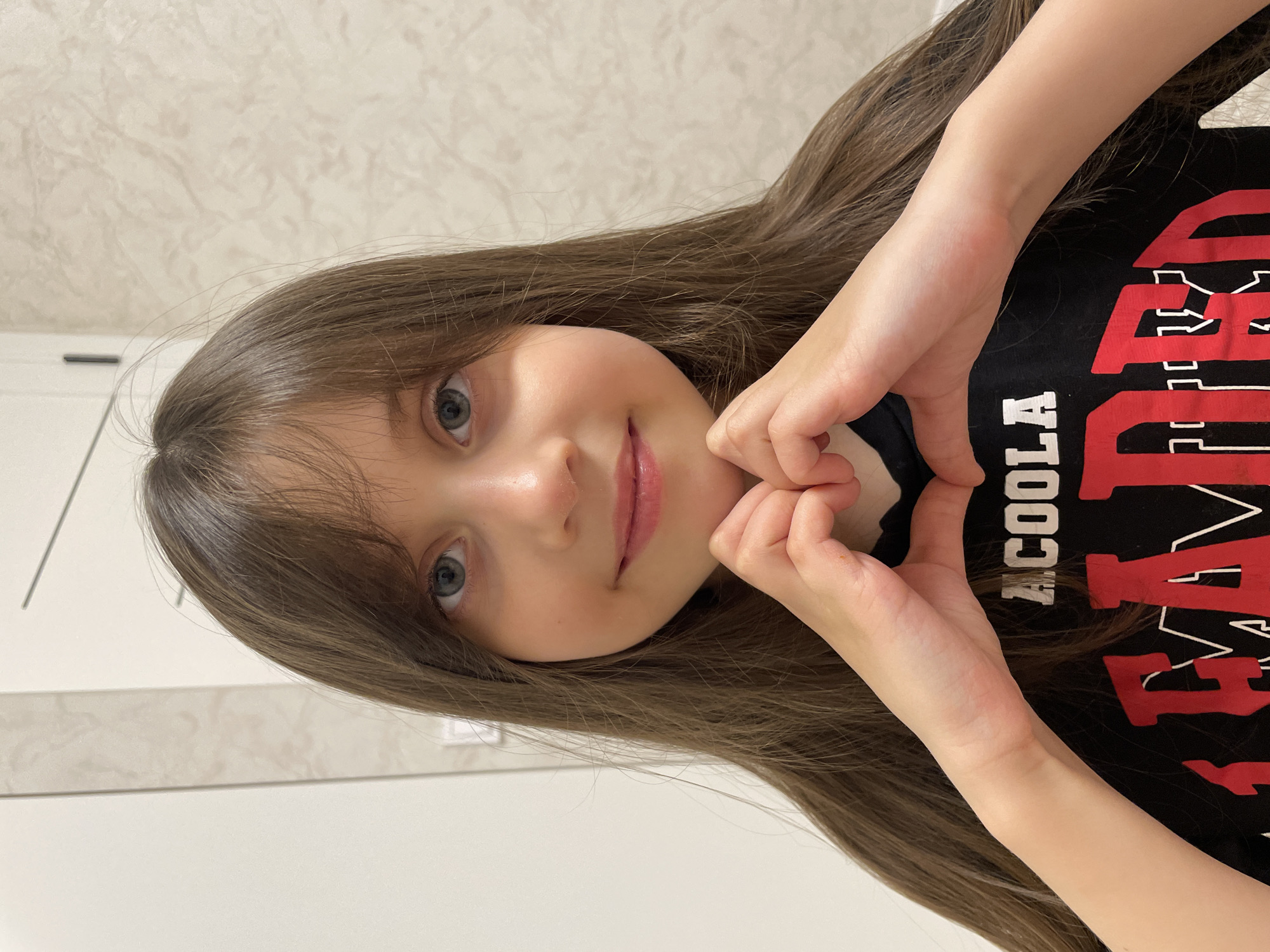 A young girl with long brown hair and blue eyes is posing indoors. She is wearing a black t-shirt with red lettering that includes the word "ACOOL". The girl is smiling slightly and forming a heart shape with her hands in front of her chest. The background consists of a light-colored wall. The location and city of Izhevsk, Russia, cannot be determined from the image.