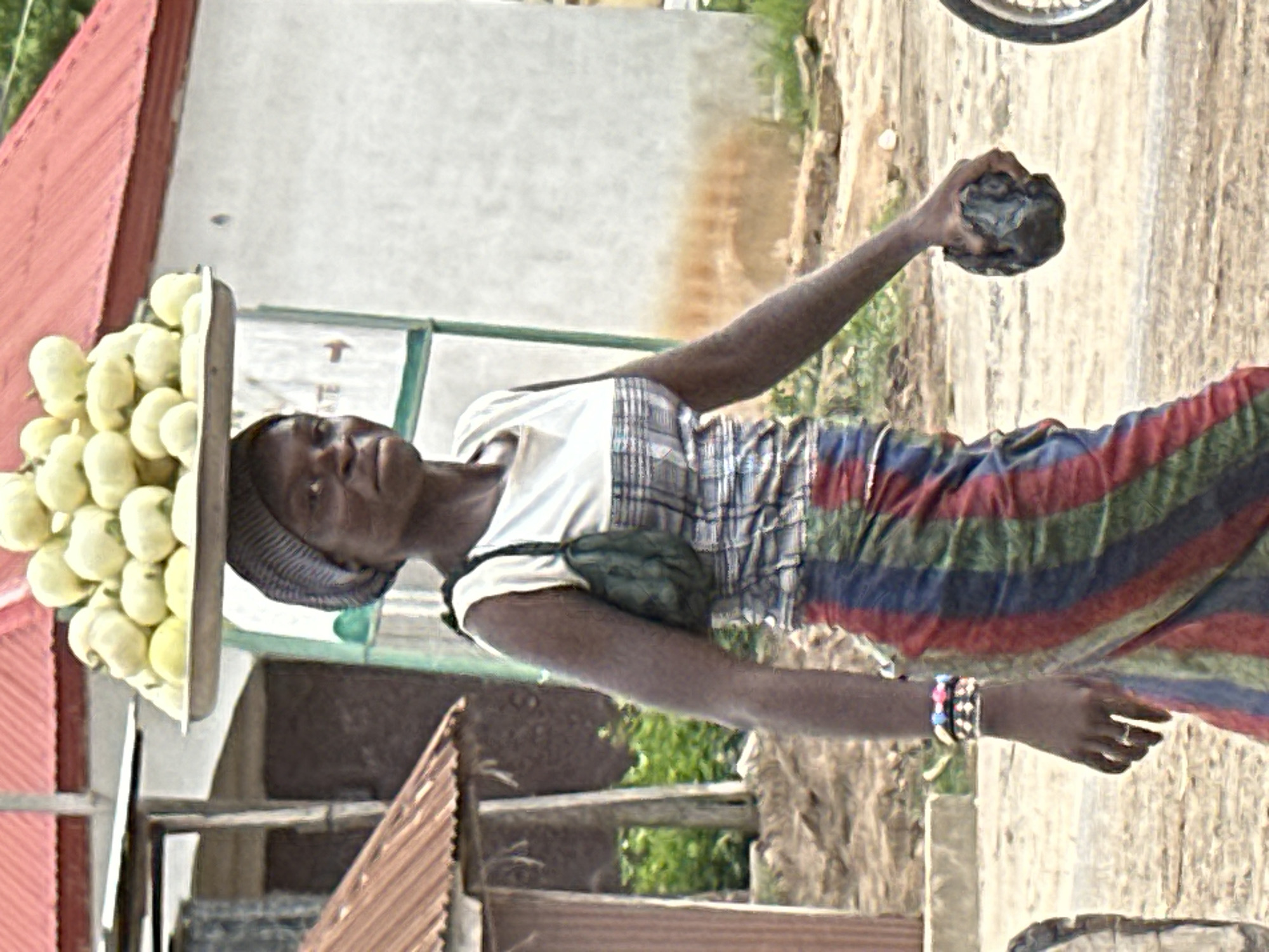 A young woman walks outdoors in Mararaba, Nigeria, carrying a tray of pale green gourds on her head. She wears a gray and white checkered top, a colorful striped skirt, and a headscarf. A small green purse is slung over her left shoulder. She has several bracelets on her left wrist. In her right hand, she holds a dark, rough-textured object, possibly a stone or lump of charcoal. The background shows buildings with red roofs, some vegetation, and a dirt road. A bicycle wheel is partially visible on the right side of the frame.