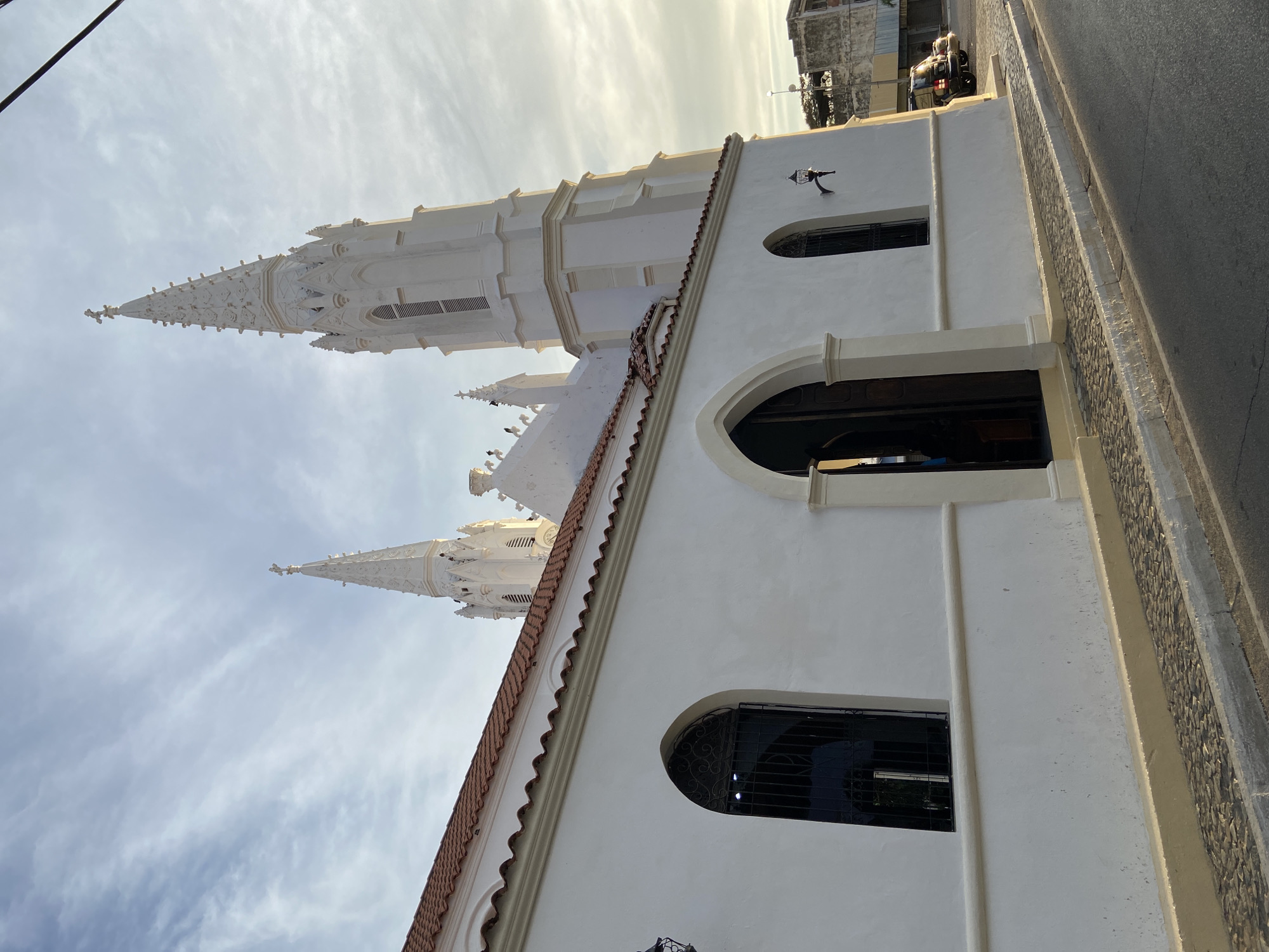 The image shows a view of the Iglesia de San Juan Evangelista in Juan Griego, Venezuela, from a low angle. The church is characterized by its white exterior and a tall, elaborate steeple. The facade of the building is visible, featuring arched windows and a red-tiled roof. A street and some cars are present along the side of the church. The sky is partially cloudy.