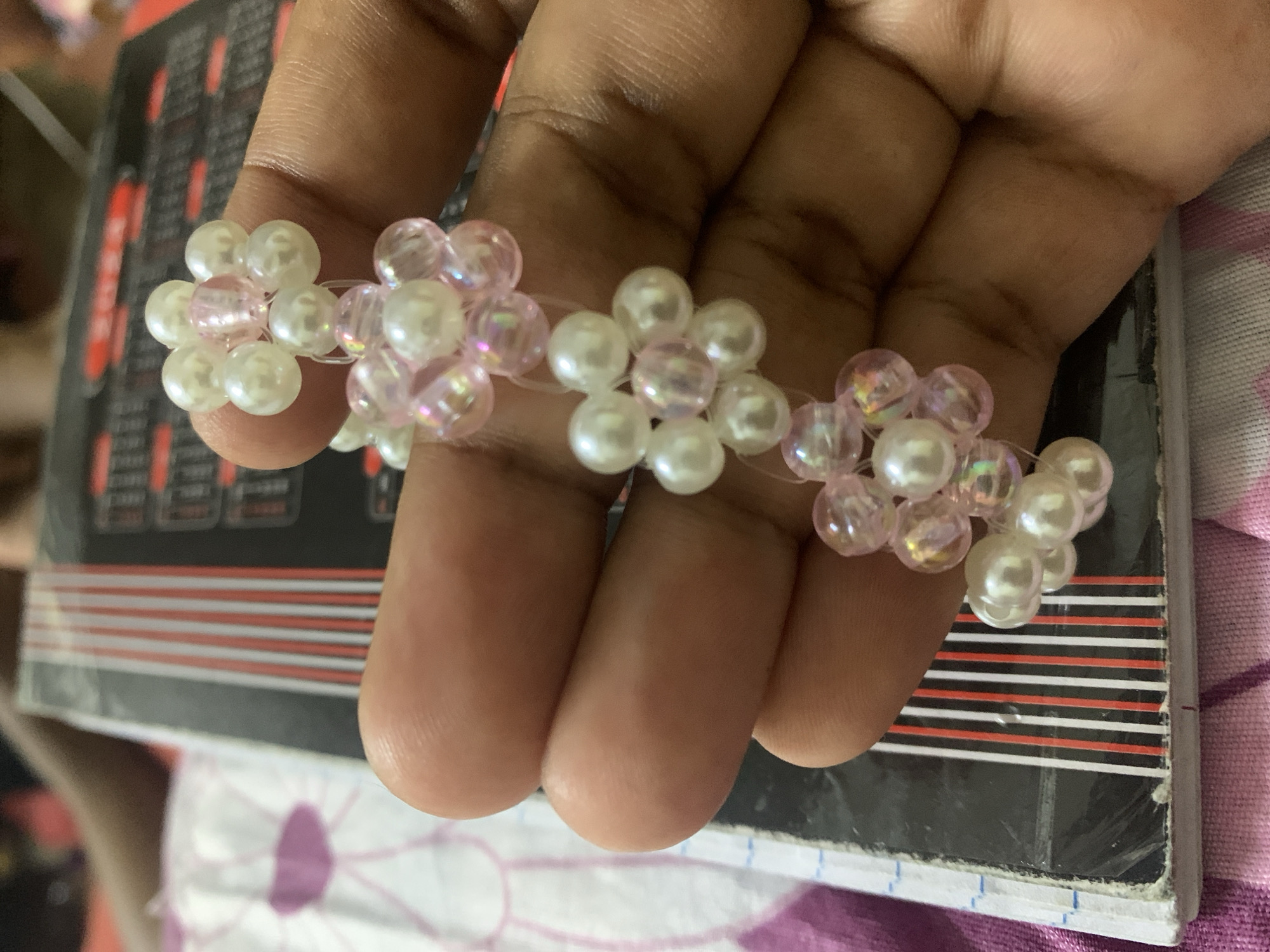 A hand holds a beaded bracelet with white and pink pearl-like beads arranged in floral patterns. The hand is set against a background of a book with black, white, and red stripes and a patterned fabric. The location is not discernible from the image.