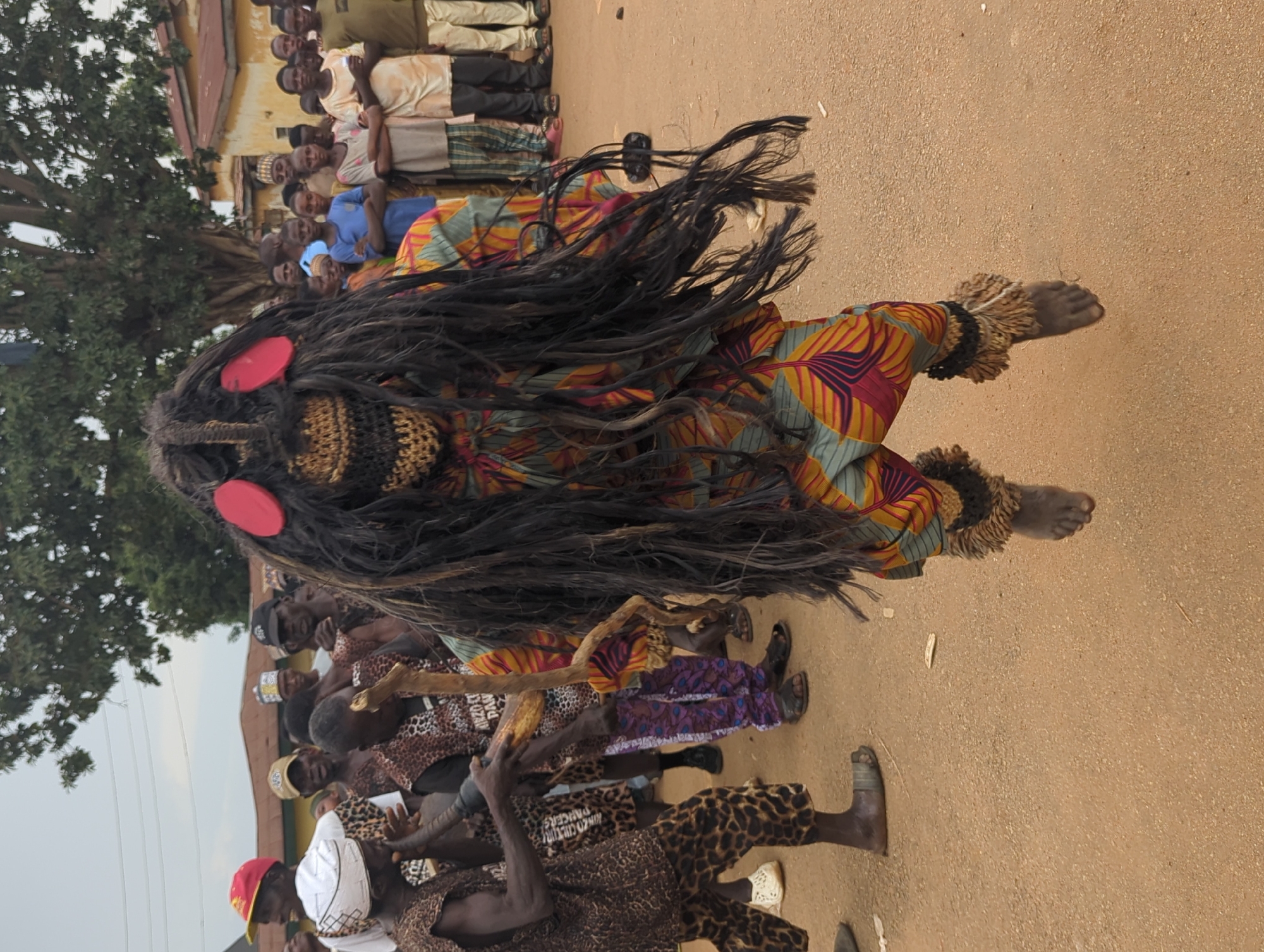 In Kafanchan, Nigeria, a procession unfolds. A figure in a vibrant, patterned costume and a mask with long, dark raffia-like strands is the focal point. The mask features two prominent red discs above a textured woven section. The figure is barefoot, adorned with anklets of a similar material to the mask's raffia, and appears to be in motion.
A crowd of onlookers lines the periphery, some wearing patterned clothing, others in Western-style attire. Some individuals carry objects like wooden poles. The ground is a dirt or sand surface. The backdrop includes trees and indistinct buildings.