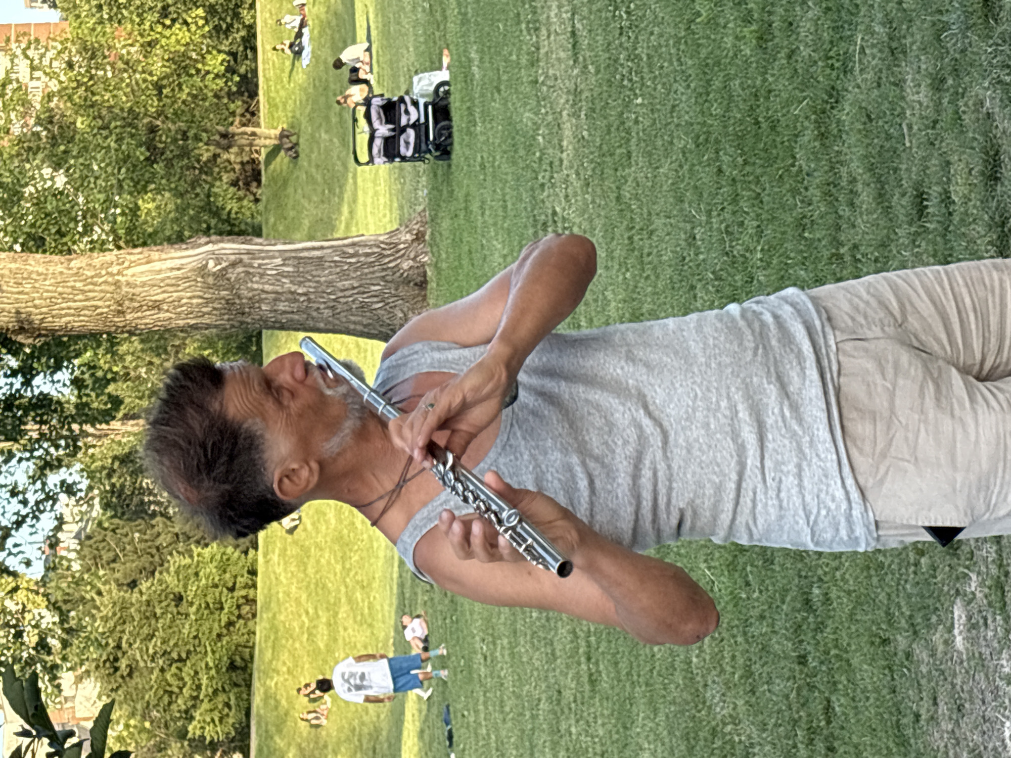 In the city of Sofia, Bulgaria, a man plays a flute in a park. He is wearing a grey tank top and tan shorts. The man has a beard and his hair is styled in a spiky manner. In the background, other people are seen relaxing on the grass, and a stroller is parked nearby. The park is green and lush with trees in the background. The sun is shining, creating a pleasant atmosphere.