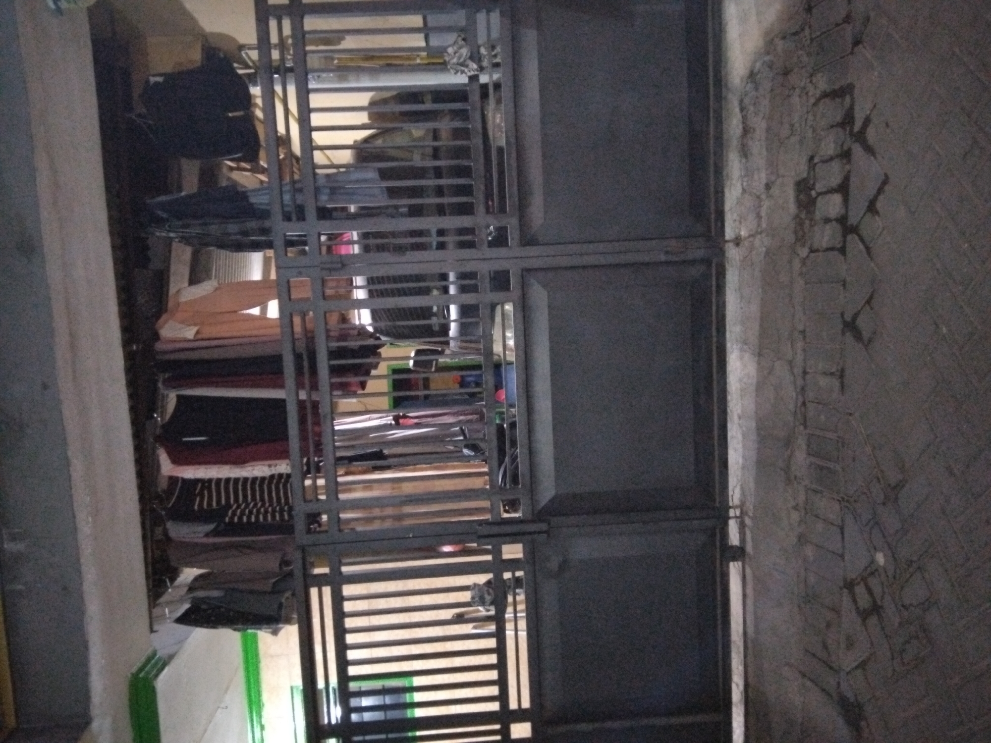 The image captures a dark, exterior view of what appears to be a residential property in Kepuh Kiriman, Indonesia. In the foreground, a grey metal gate with vertical bars and a solid lower section blocks access. The gate is closed and appears to be made of wrought iron or a similar metal. Behind the gate, clothes are hung out to dry on a clothesline suspended from a covered structure. The clothes consist of various garments, including shirts, pants, and possibly towels, in a range of colors and patterns. Some of the clothes are dark, while others are lighter, and one garment appears to be orange. Further behind the clothesline, a large, dark-colored object, possibly an air conditioning unit or a piece of machinery, is visible. The background shows a wall or building structure with some yellow and green accents. The ground in front of the gate is paved with dark, patterned tiles, some of which are cracked or uneven. The lighting in the scene is dim, suggesting it might be evening or nighttime, with artificial light illuminating some areas. There are no people visible in the image.