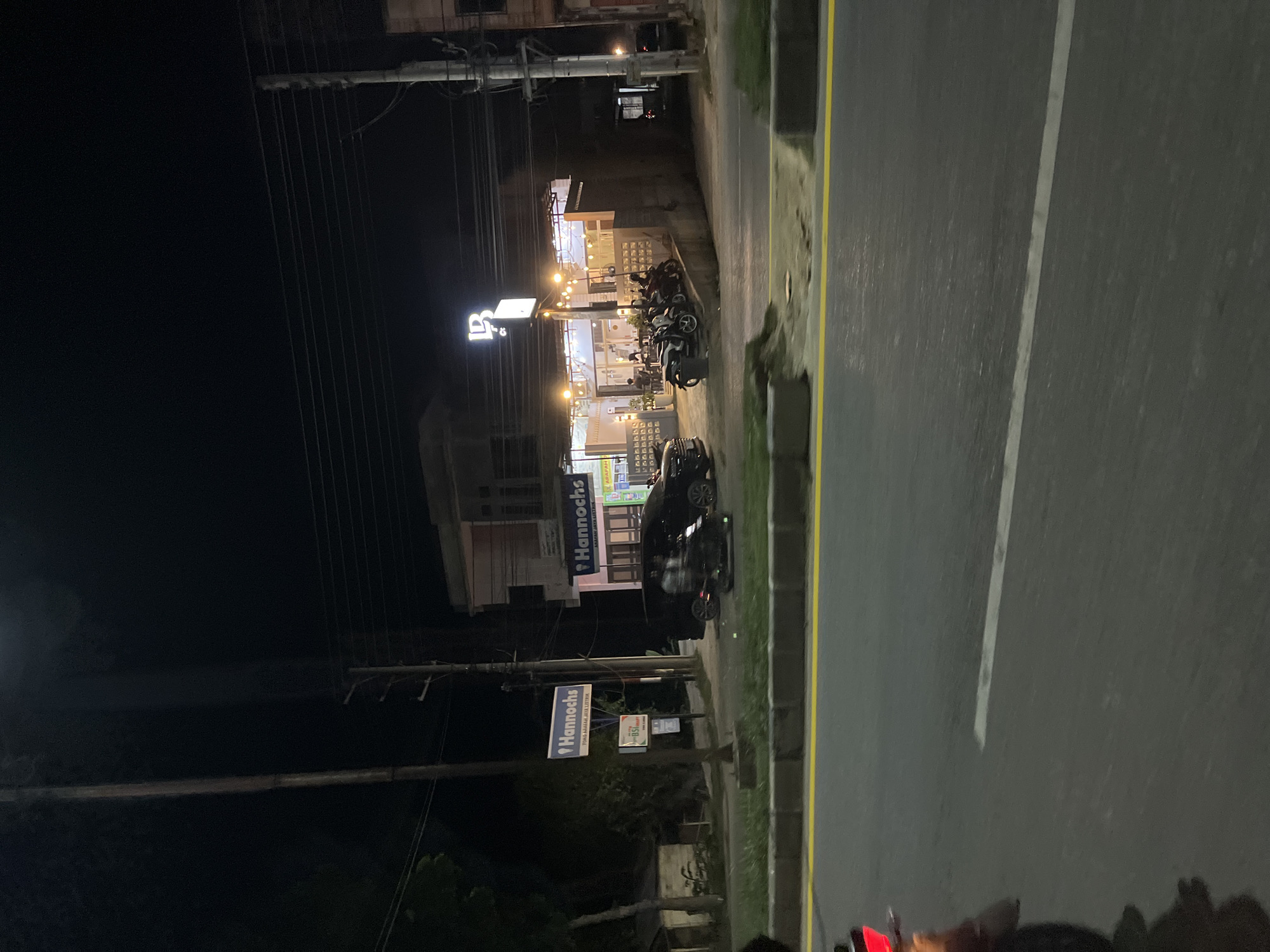 This is a nighttime exterior shot taken from a moving vehicle on a road in Bundar, Indonesia. The scene features a street with a yellow painted line and a white line, indicating traffic lanes. Across the road, several businesses are visible, illuminated by artificial lights. One building has a prominent sign that reads "Hannochs" with the tagline "TOKO ALAT-ALAT LISTRIK" (Electrical Equipment Store). Adjacent to it, another "Hannochs" sign is visible. Further down, a building with outdoor lighting and people sitting at tables suggests a cafe or restaurant, with a sign bearing the letters "LB" and "Co." Several motorcycles are parked outside this establishment. Black vehicles, including a car and what appears to be a scooter or motorcycle, are also present on the scene. Utility poles and power lines are visible against the dark sky. The overall impression is of a commercial area at night.