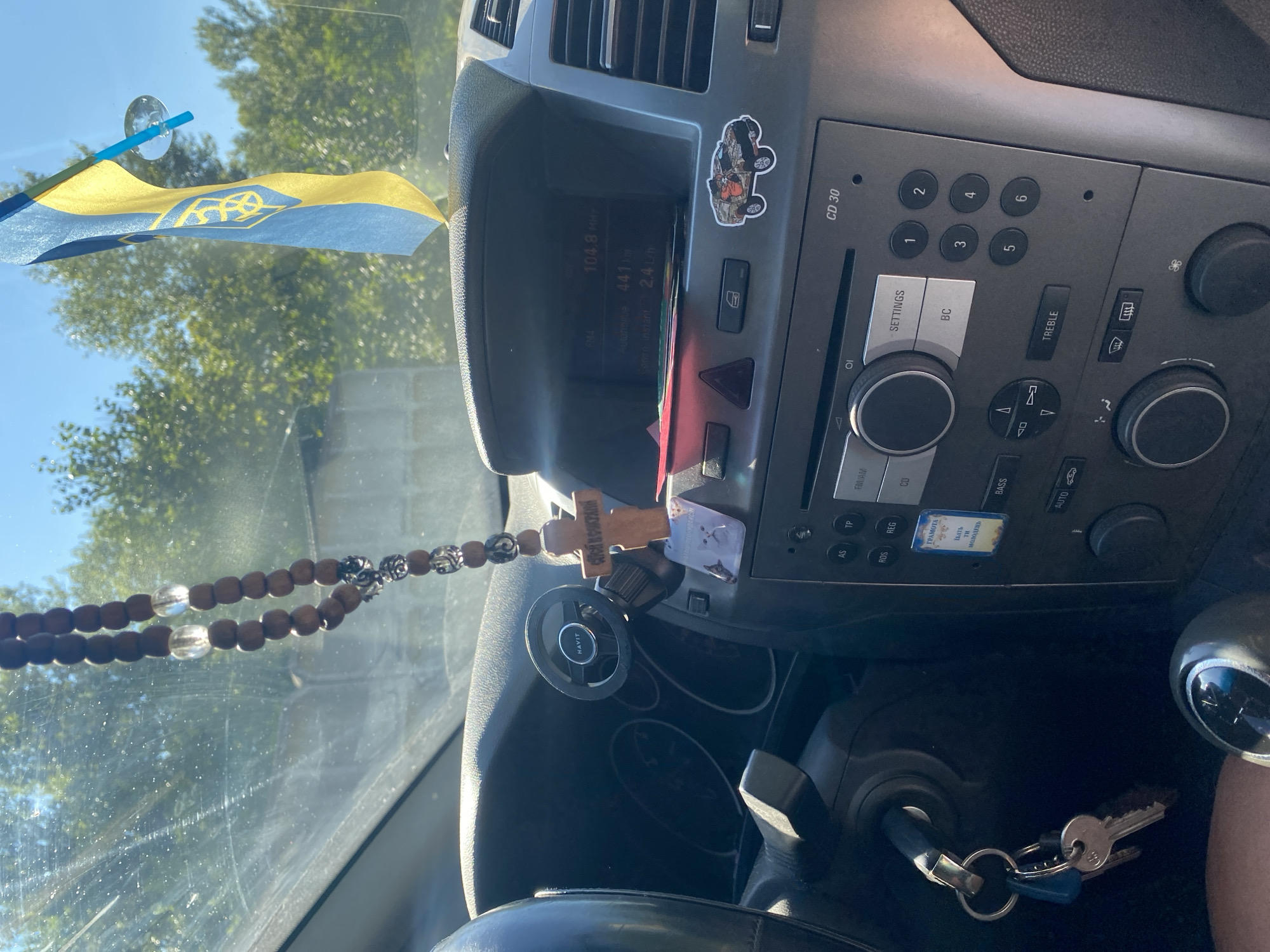 The image shows the interior of a car. On the dashboard, there is a car stereo with a display screen showing "104.8 MHz", "441 km", and "2.4 L/h". A wooden rosary with silver accents and clear beads hangs from the rearview mirror. A small wooden cross with an inscription is attached to the rosary, along with two small stickers, one of a cat and another with an inscription. A Ukrainian flag is hanging from the windshield. The steering wheel and car keys are visible on the left side of the image. The car appears to be parked outdoors on a sunny day, with trees and a building visible in the background. The location is the city of Dnipro, Ukraine.