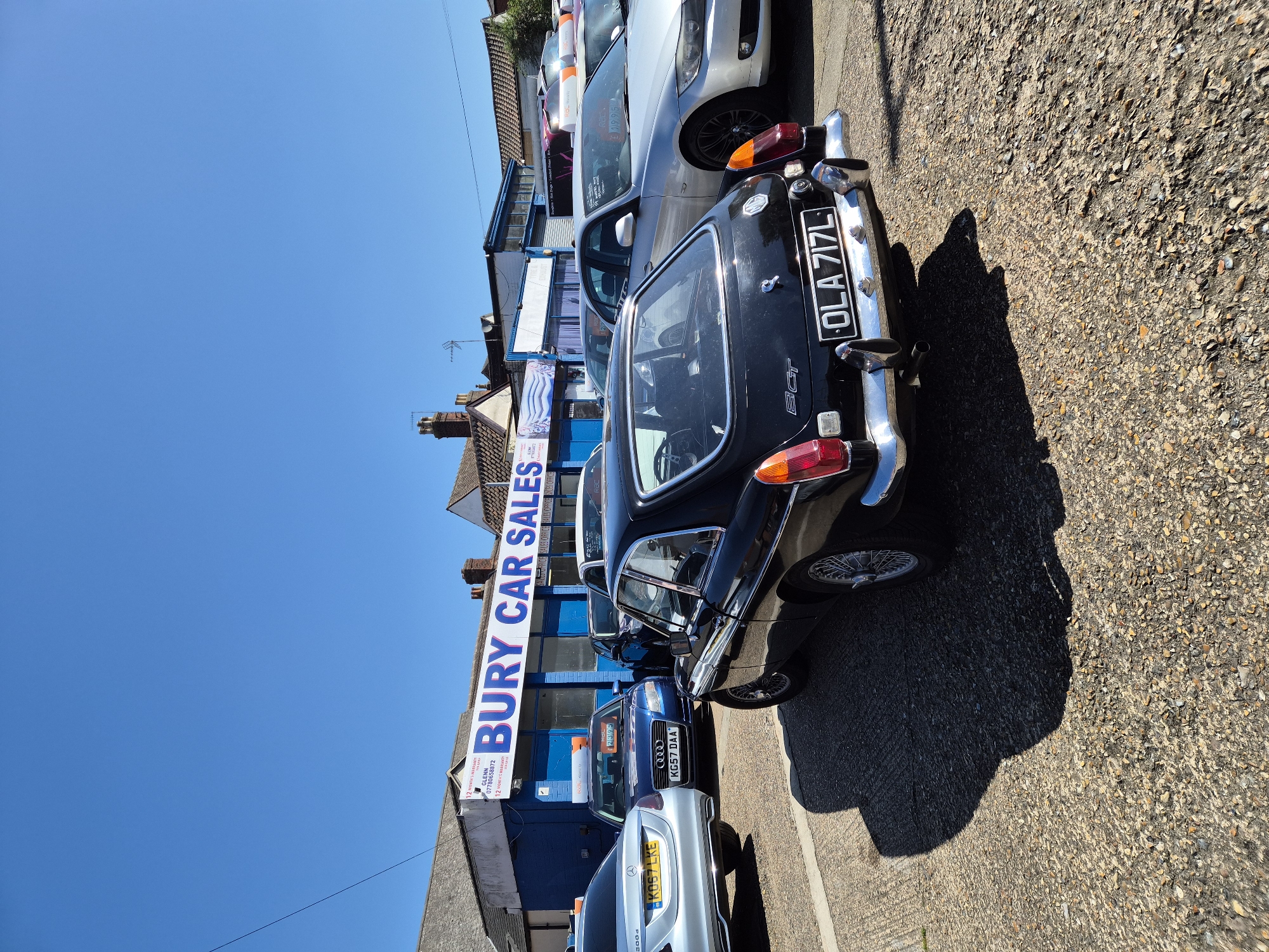 An outdoor, eye-level photograph, rotated 90 degrees clockwise, captures a car sales lot on a bright, sunny day, likely in West Suffolk, United Kingdom. The clear blue sky dominates the upper part of the frame, indicating midday or early afternoon conditions with strong shadows cast on the gravel ground in the foreground.
The primary subjects are several cars parked along a roadside, adjacent to a commercial building identified as a car dealership. In the foreground, a dark-colored classic MGB GT, distinguishable by its "GT" badge and chrome bumpers, displays the British license plate "OLA 717L". Behind it, a blue Audi with license plate "KG57 DAA" is visible, featuring a price sticker with "£2395". Further down the line, a silver Mercedes, bearing the license plate "KO57 LKE", is partially visible. Other vehicles, including a silver BMW and another car with a red element, are parked further along the street, suggesting an active car sales business.
The dealership building features a large, prominent blue and white sign stating "BURY CAR SALES". Below this, smaller text advertises "12 MONTHS WARRANTY", a contact name "GLENN", and a phone number "07780658872". The building's windows display repeated text "SALE OFFICE". No people are visible in the image. The scene suggests a typical commercial setting for car sales on a pleasant day.