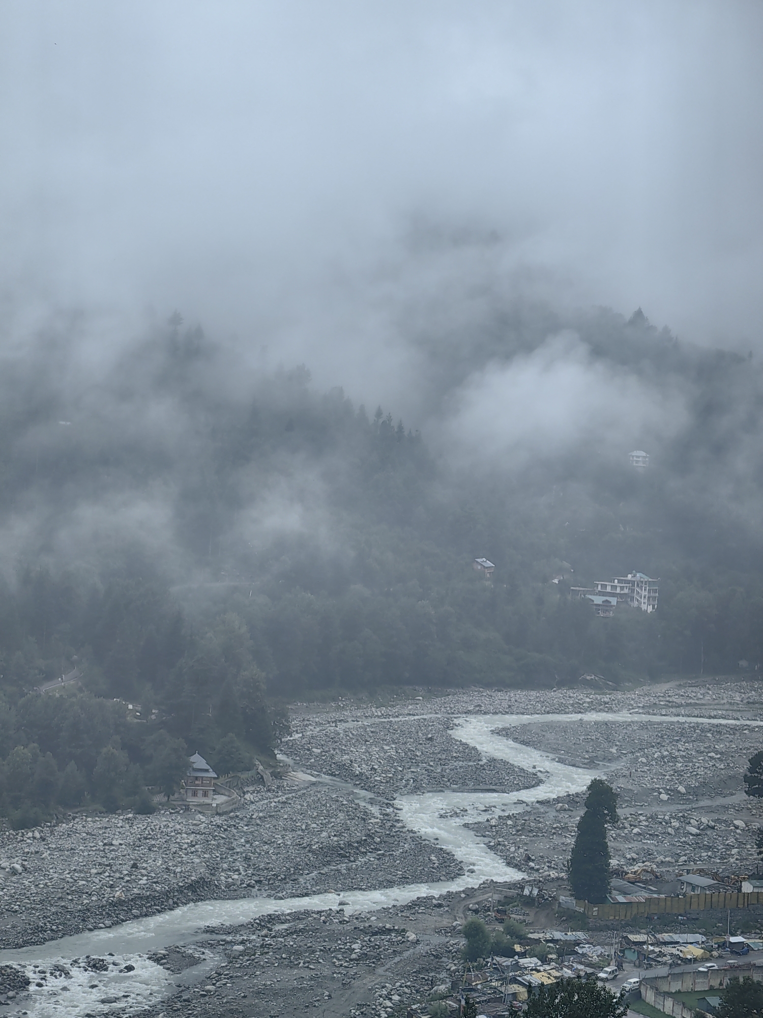 This is a wide shot of a misty, mountainous landscape. In the foreground, a river flows through a wide, stony riverbed. On the left bank of the river, a small, ornate building with a dark roof sits atop a stone wall. The river then curves to the right, flowing towards the bottom right of the image. Beyond the riverbed, a dense, green forest covers the mountainside, with a few buildings interspersed among the trees. White clouds and mist obscure the upper slopes of the mountains, giving the scene a surreal and atmospheric quality. The overall impression is one of natural beauty and tranquility, with a touch of human habitation.