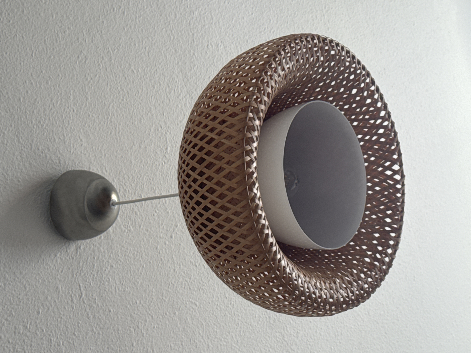 A wall-mounted light fixture is shown on a textured white wall. The fixture consists of a round, metal base attached to the wall with a thin rod extending to a brown, woven, donut-shaped lampshade. The inner part of the shade contains a gray cylindrical component, presumably housing the lightbulb. The scene is indoors, location Unknown, Unknown.