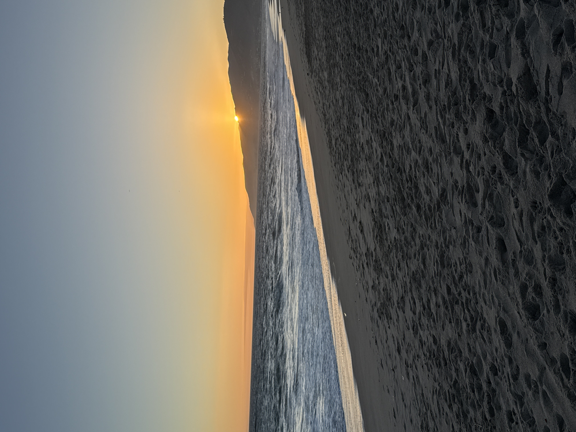 The image depicts a serene sunset scene at a beach in Malibu, California. The sky transitions from a pale blue at the top to a warm orange glow near the horizon, where the sun is partially obscured by a distant, silhouetted mountain range. The ocean stretches across the middle ground, with waves gently rolling towards the shore. The water reflects the golden hues of the sunset, creating a shimmering path across its surface. The foreground is dominated by dark, sandy beach, marked with numerous footprints, suggesting recent human activity. The overall atmosphere is tranquil and picturesque.