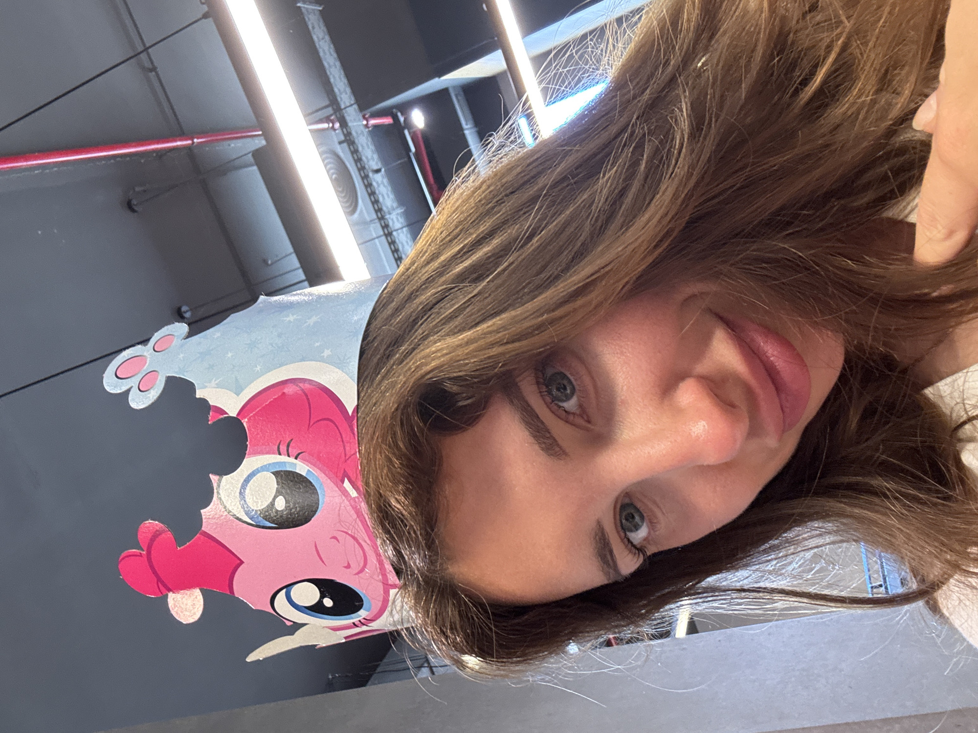 A tilted close-up shot shows a woman with light brown hair and blue-green eyes, posing next to a cartoon drawing of Pinkie Pie from My Little Pony. The background is an out-of-focus industrial-style ceiling with exposed pipes and fluorescent lighting. The location is in the city of Sueca, Spain.