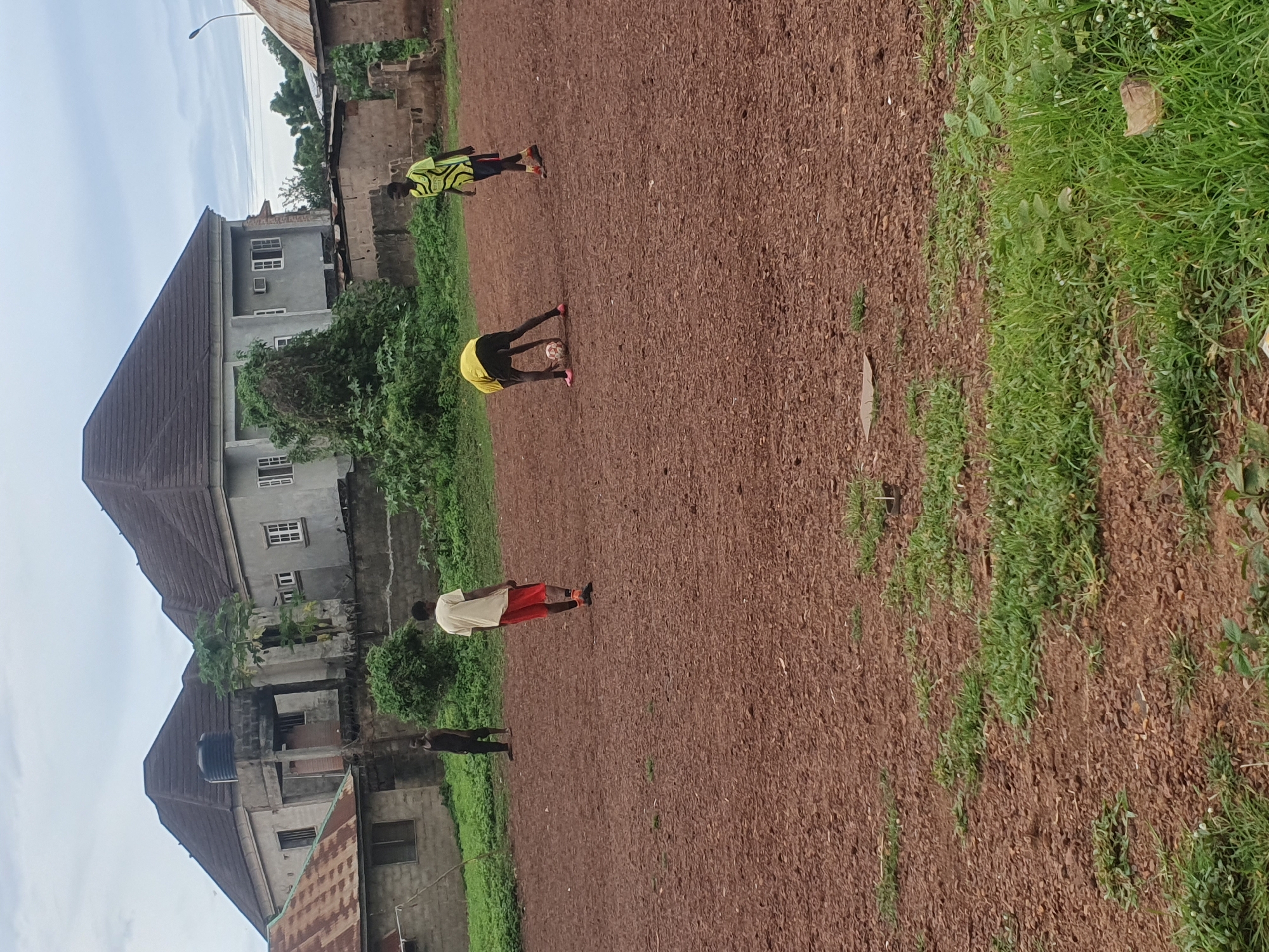 The image depicts an outdoor scene in Idah, Nigeria, featuring four young men on a reddish-brown dirt and gravel field with scattered patches of green grass. In the mid-ground, a man in a yellow and black patterned long-sleeved shirt and dark shorts bends over a white and black soccer ball, wearing pink athletic shoes. Another man, similarly dressed in a yellow and black patterned shirt and dark shorts with orange athletic shoes, stands upright to his right. Further left, a man in a light t-shirt and red shorts stands with his back to the viewer, and a fourth man in a dark tank top and dark shorts stands to the far left. The background consists of residential buildings under an overcast sky. On the left, a single-story building with a rusted corrugated metal roof and concrete walls is visible. In the center, a prominent two-story concrete building with a dark, multi-hip roof, multiple white-framed windows, and a black water tank on a smaller adjacent roof structure is present. To the right, another single-story building with a rusted roof and a distant street light pole are visible. Green vegetation, including bushes and trees, grows around the buildings and along the edges of the field.