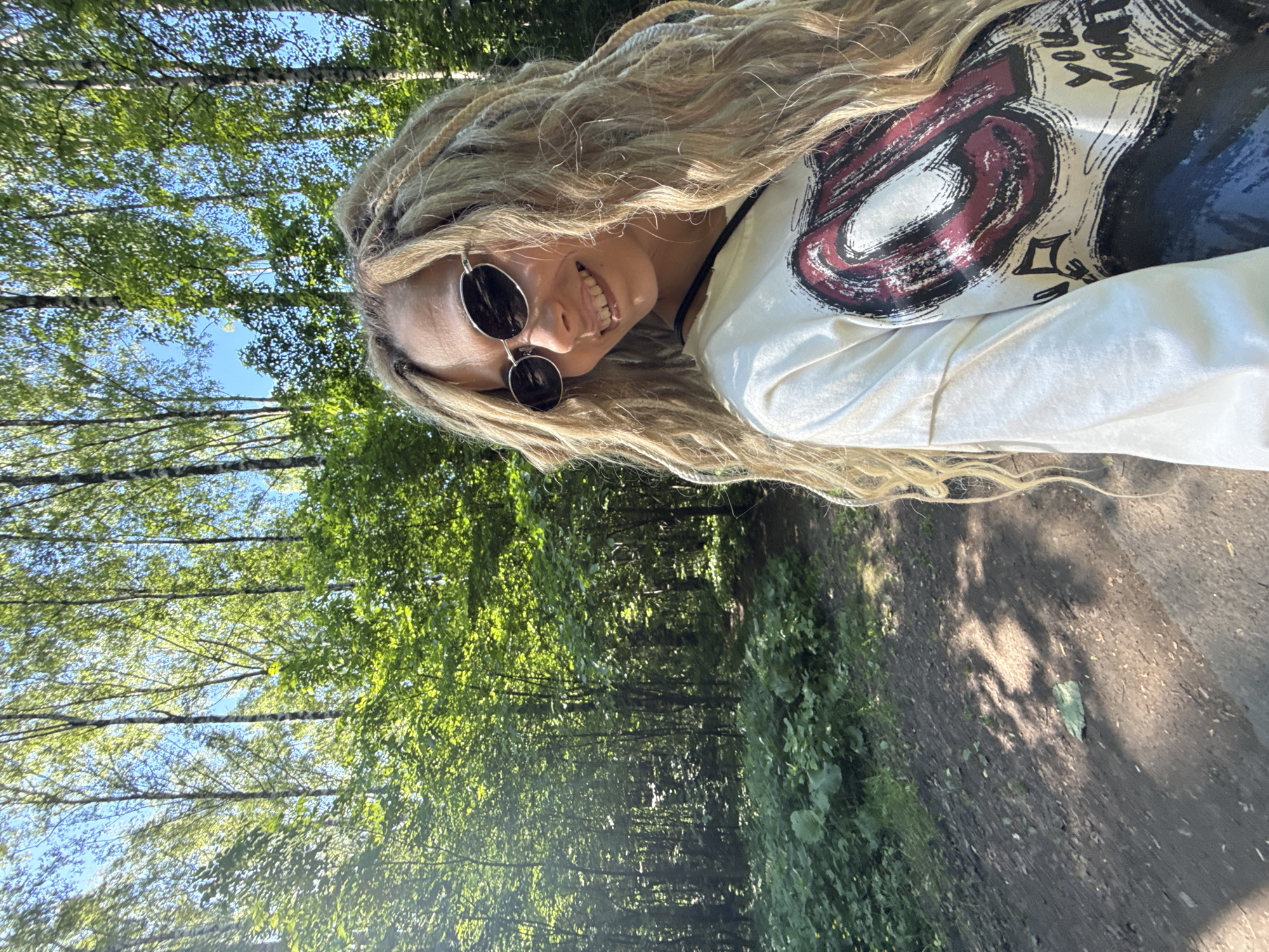 A female individual is captured in a close-up, waist-up portrait within a dense wooded area in Rusanovo, Russia. She has long, blonde dreadlocks and wavy hair cascading over her shoulders. She wears dark, oval-shaped sunglasses with silver frames and an off-white, long-sleeved t-shirt featuring a large, stylized red and black graphic design with partially visible text, "you want". A black strap is visible across her left shoulder. The subject is smiling, revealing her upper teeth, and is positioned on the right side of the frame, looking back towards the camera over her left shoulder.
The background consists of a sunlit deciduous forest under a clear blue sky. Tall trees with slender trunks and vibrant green foliage dominate the upper and mid-ground. A dirt path is visible in the lower-left and central portions of the image, winding into the distance, bordered by thick green undergrowth, including large-leafed plants. Dappled sunlight filters through the tree canopy, creating distinct patterns of light and shadow on the path and vegetation.