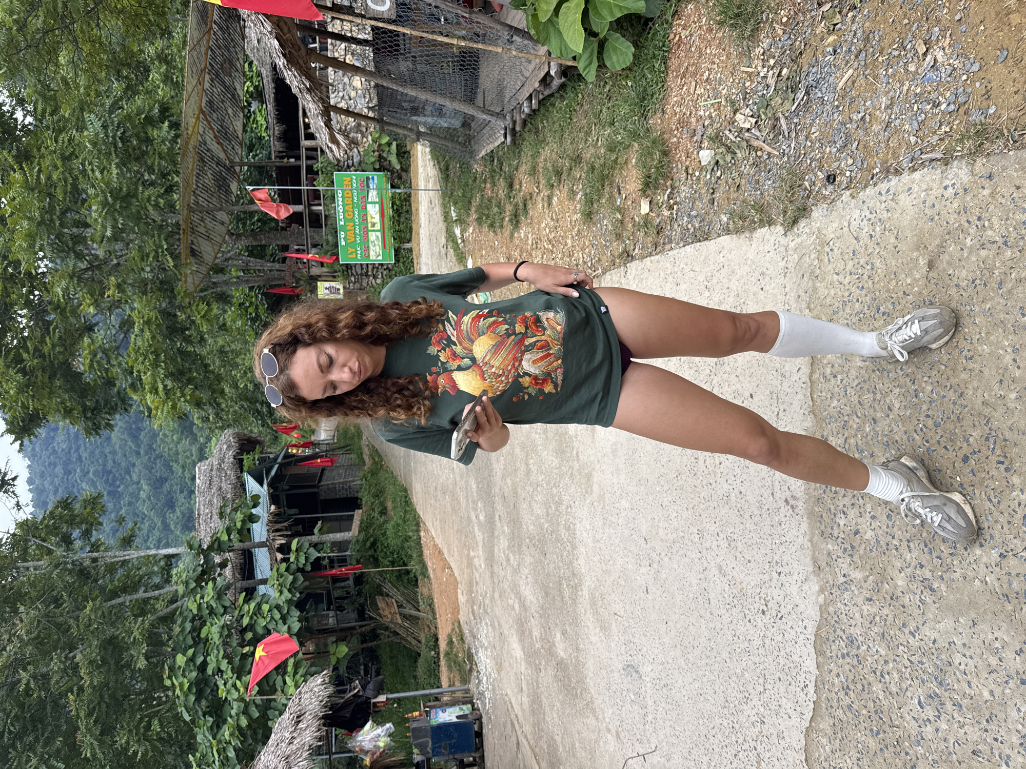 A woman with curly, brown hair stands on a paved path, looking down at her phone. She is wearing a dark green t-shirt with a colorful rooster graphic, dark red shorts, white socks, and gray sneakers. Behind her, the path leads past several thatched-roof buildings and lush green trees. Red flags with yellow stars are visible, some attached to poles and others hanging from structures. A green sign with Vietnamese text is posted on a pole. The environment appears to be a rural area, possibly a village or tourist spot, with a mountainous backdrop covered in dense forest. The scene is outdoors, with natural light illuminating the area.
