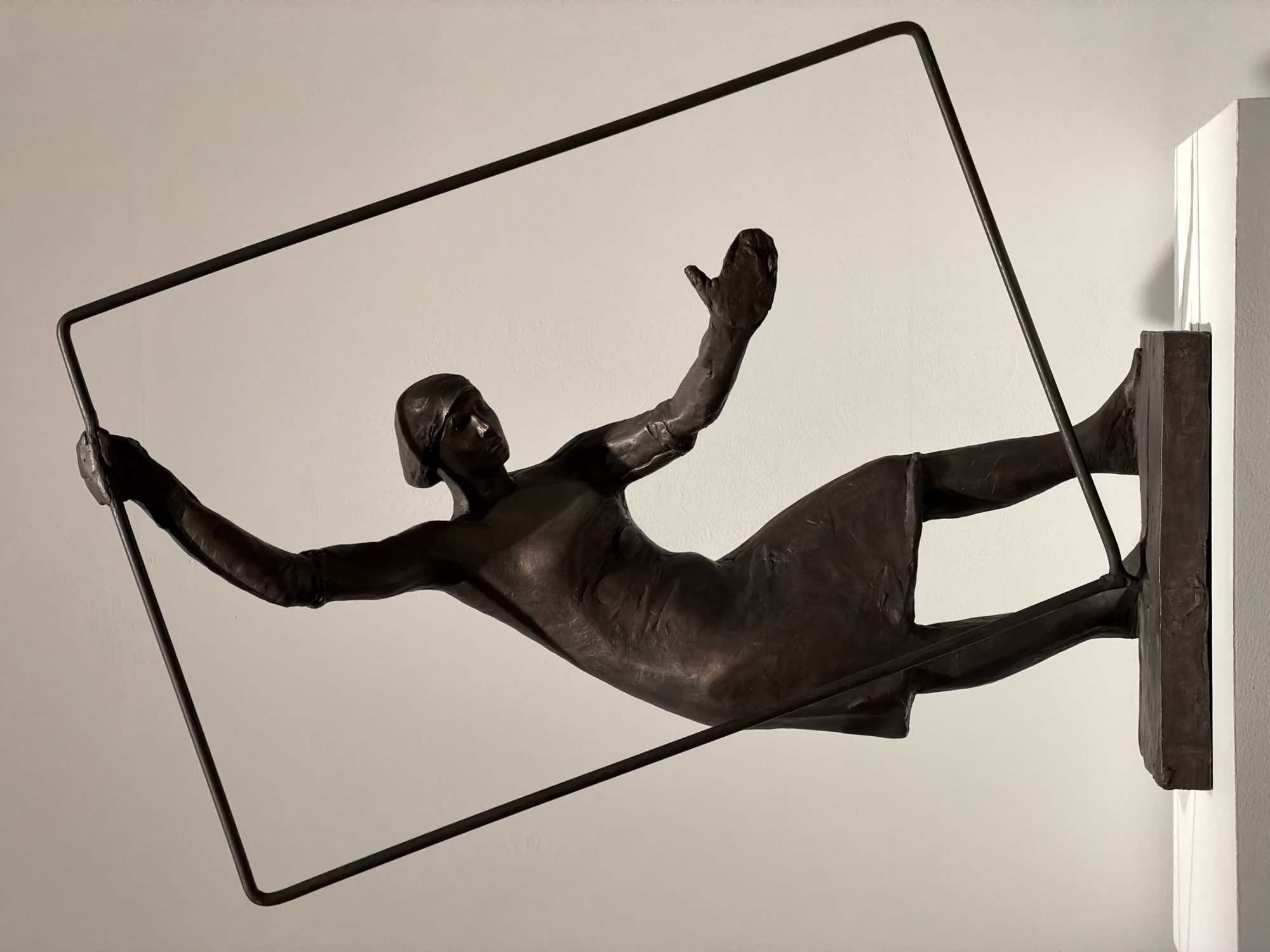 A bronze statue of a woman wearing a head covering is affixed to a white wall. She is positioned horizontally, leaning towards the viewer. The woman's left arm extends out holding part of a rectangular frame, while her right arm is raised up. Her legs are also extended, with the right leg attached to a bronze base affixed to the wall. The background is a smooth, light-colored wall.