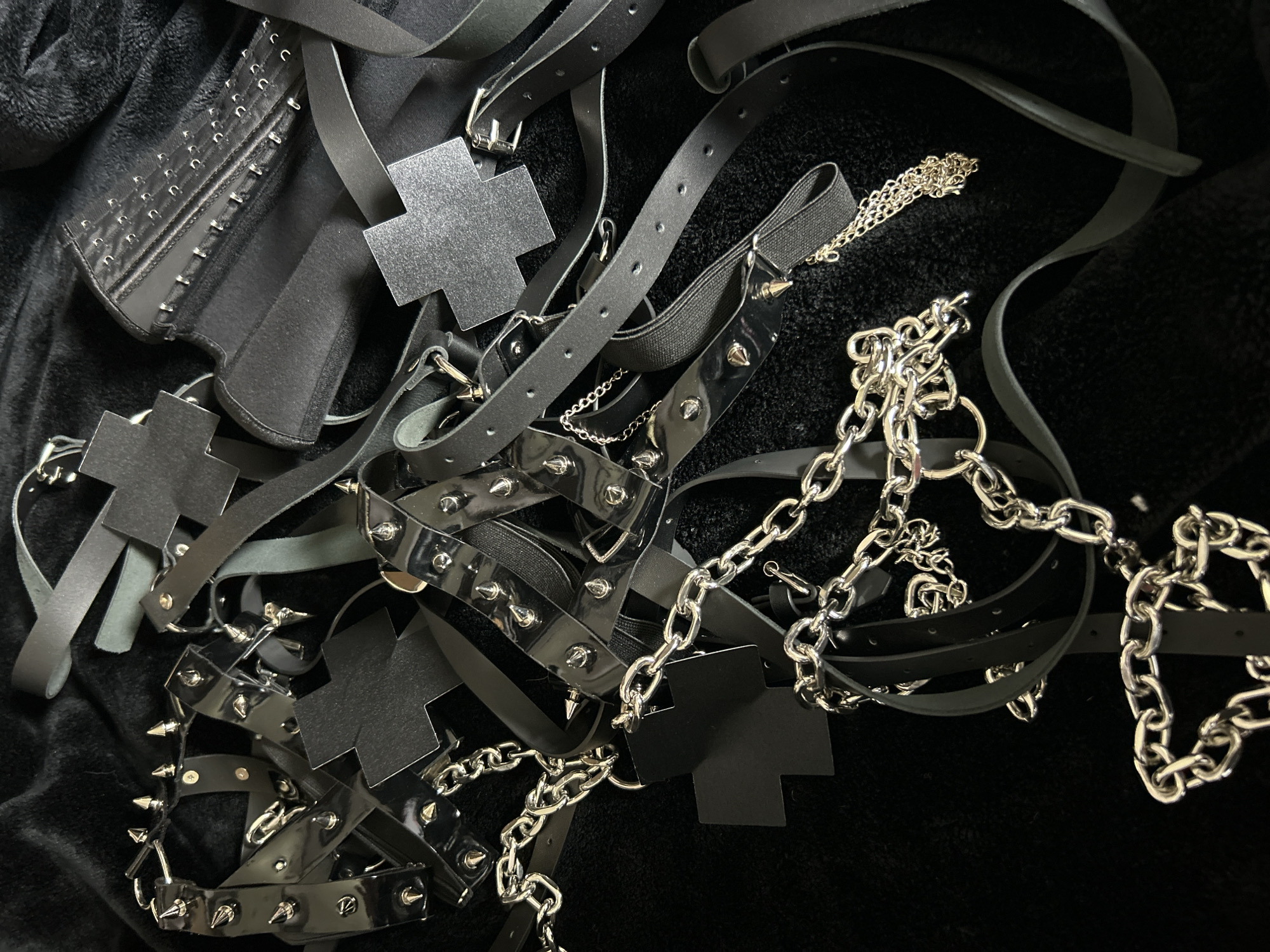 The image depicts a collection of dark-colored, alternative fashion accessories scattered on a textured, dark surface. The accessories include a black corset, multiple black leather straps, some adorned with metal studs and cross-shaped appliques, and silver-colored metal chains. The scene appears to be indoors, illuminated by artificial light. There are no people visible. There is no information to suggest the location is Cape Coral, United States of America.