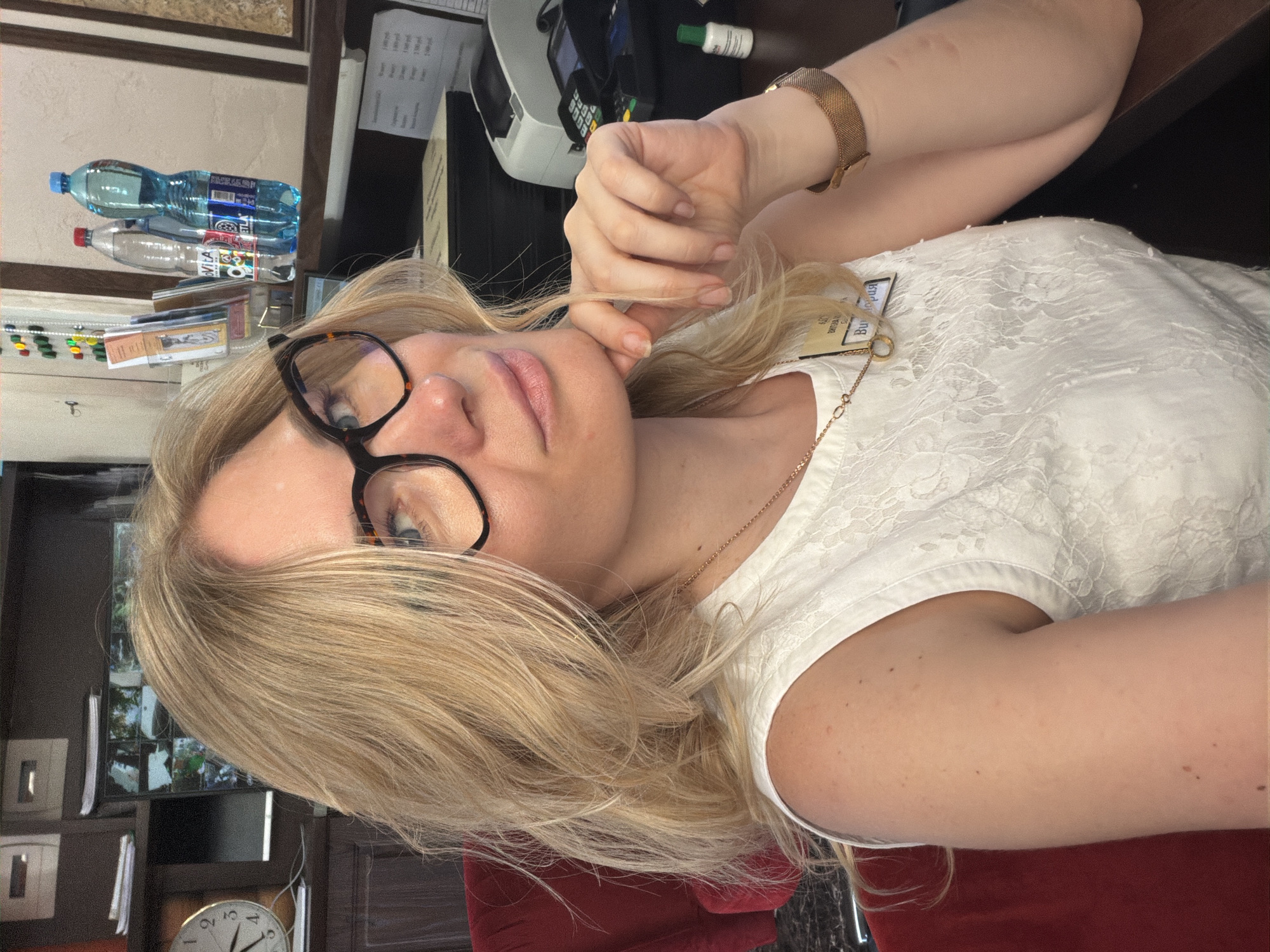 A woman with blonde hair and glasses is taking a selfie in an indoor setting, possibly an office or reception area. She is wearing a white lace top and a gold necklace. Her right hand is resting on her chin, and she is wearing a gold watch on her right wrist. In the background, there are shelves with bottles of water, a clock, and some office equipment. The scene is dimly lit, with a focus on the woman. There is no indication of the city of Sochi, Russia.