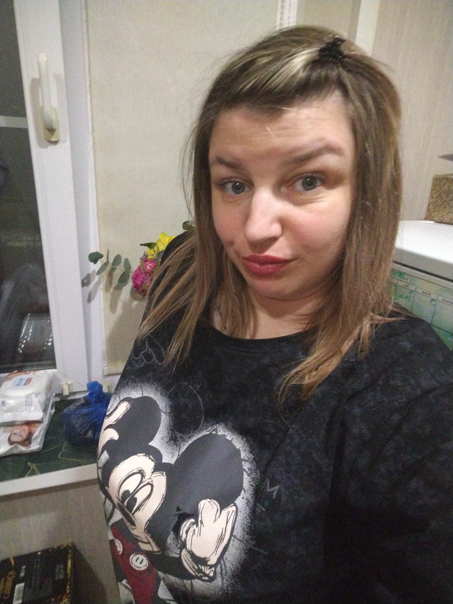 A woman is shown in a selfie indoors. She has fair skin, light brown hair with blonde highlights, and is wearing a black T-shirt with a Mickey Mouse design. The background is a room setting with a window, which appears to have some items on the windowsill, including packages of wet wipes and a dark blue bag. There are also flowers behind the person. In the upper right corner of the frame, part of a white appliance or piece of furniture is visible with what appears to be a chart or schedule on it. The image was likely taken indoors in a residential setting, but without more context, I cannot verify the location in the city of Yaroslavl'.