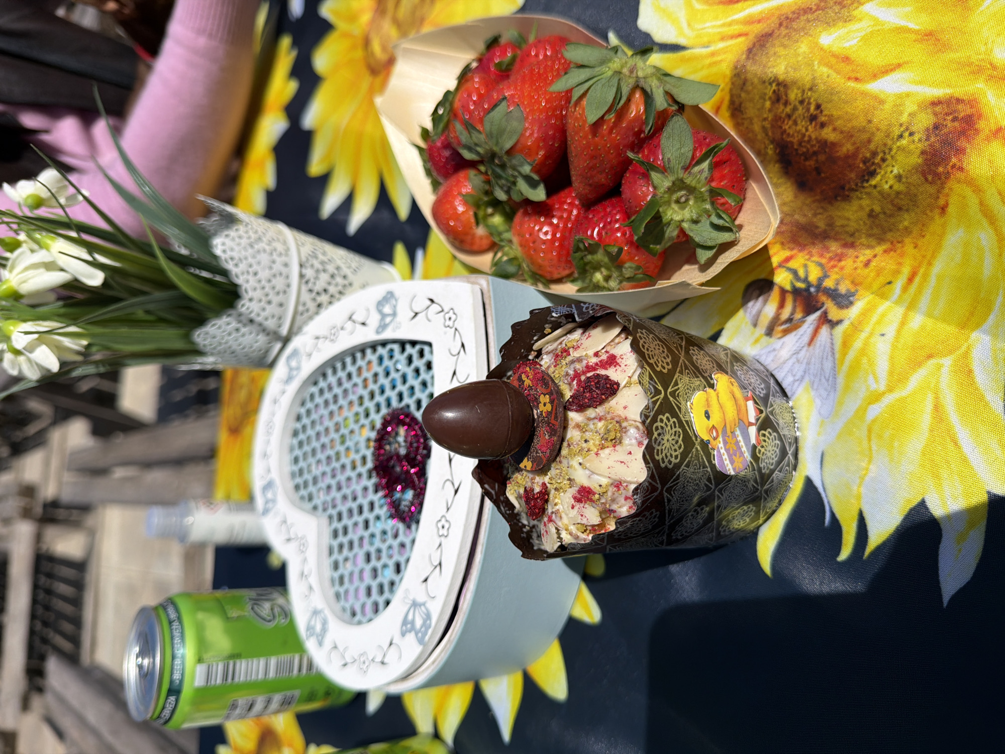 A bright and sunny outdoor scene captures a festive arrangement of treats and decorative items laid out on a table. The table is covered with a dark blue/black tablecloth featuring a vibrant pattern of large yellow sunflowers and bees.
In the foreground, a decorative cupcake or muffin is prominently displayed. It's encased in a dark brown and gold patterned paper wrapper, adorned with a yellow Easter chick sticker. The cupcake is topped with a generous swirl of light-colored frosting, sprinkled with red dried fruit pieces (possibly raspberries) and green pistachios. A small, dark chocolate egg rests on top, accompanied by a piece of decorated chocolate.
To the left, a heart-shaped light blue or grey decorative box is visible. Its front panel has a mesh-like design, revealing a glittery red heart inside. The box edges are decorated with delicate white floral and butterfly patterns. Next to it, a small bouquet of white flowers, resembling snowdrops, is wrapped in white paper with a lace-like texture.
Further back, a small wooden boat-shaped container is filled with fresh, ripe red strawberries, their green stems providing a vibrant contrast. Partially visible on the left are a green beverage can with a barcode and some text, possibly "WEIHSS... BIERE...", and a small clear plastic bottle. A person wearing a pink sleeve is subtly visible in the upper left corner, suggesting human presence.
The bright daylight indicates a clear, sunny day, creating a cheerful and celebratory atmosphere, possibly for an Easter gathering or picnic in an outdoor setting like Monistrol de Montserrat, Spain.