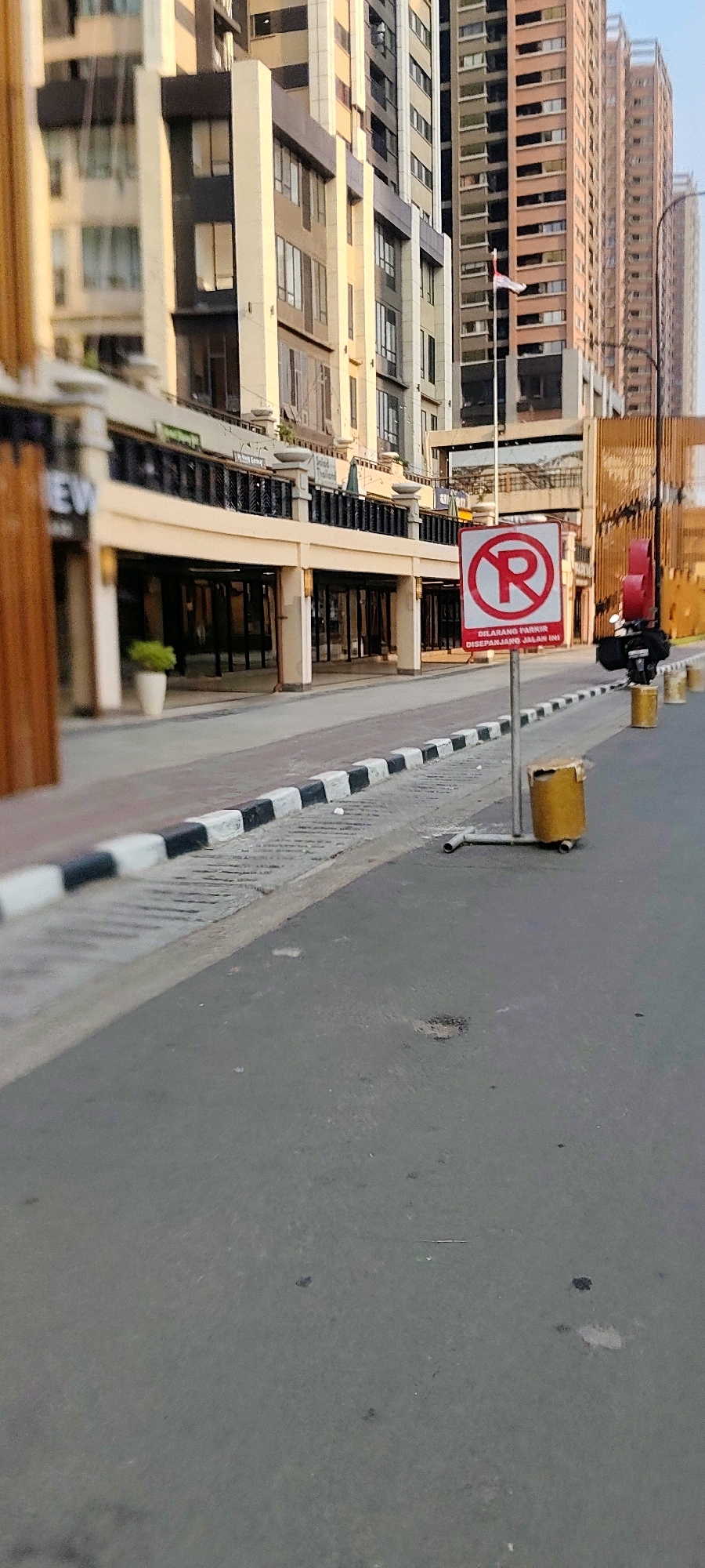 In Jayamukti, Indonesia, a street-level view captures a modern urban environment with tall buildings. A prominent red and white "No Parking" sign with text in Indonesian is positioned on a metal stand in the foreground. Adjacent to the sign is a yellow trash receptacle, partially damaged. The street is paved, with a distinct black and white striped curb lining the pedestrian walkway. Behind the curb, the walkway features a textured surface and what appear to be concrete bollards. The background is dominated by multi-story buildings, characterized by their contemporary architecture, large windows, and varying facades. A flag is visible on a pole between two buildings. The lighting suggests it is daytime, possibly late afternoon, given the warm tones. No people are visible in the immediate vicinity of the sign or trash can.