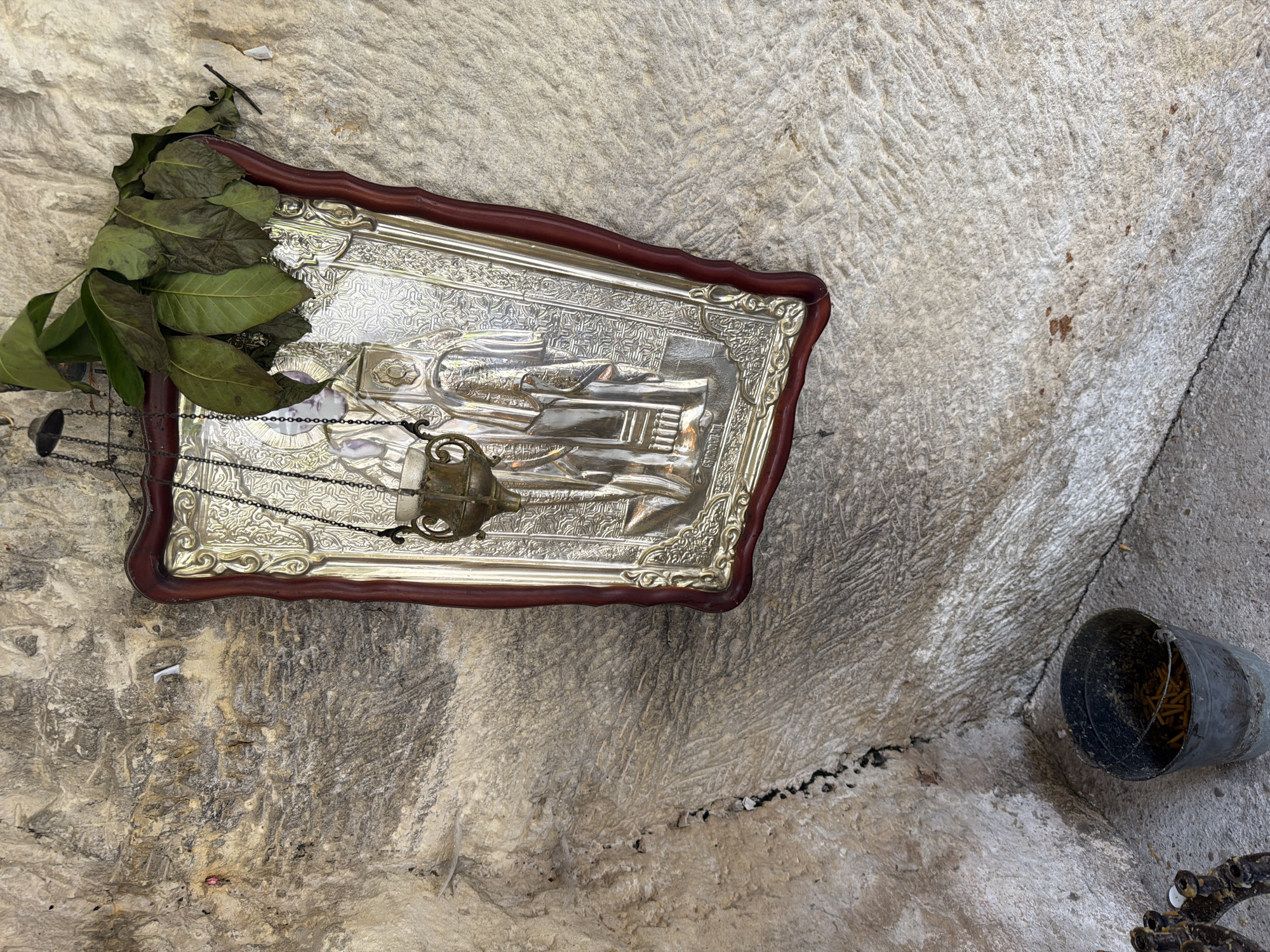 The scene depicts a devotional display set against rough-hewn stone walls and a flat floor in Basarbovo, Bulgaria. A rectangular religious icon, featuring a dark red wooden frame and a silver-plated embossed metal panel depicting figures, is affixed to the upper right section of the wall. Suspended by chains from a hook above the icon is an ornate, dark metal oil lamp or censer, positioned centrally in front of the icon. A small cluster of green leaves rests on the upper right corner of the icon, extending onto the adjacent wall surface. The walls are composed of light-colored, irregular stone with visible striations, textures, and darker fissures, particularly in the lower left quadrant. On the ground, a dark grey metal bucket containing multiple light-colored cylindrical objects, consistent with spent candle stubs, is present in the lower center. To the immediate left of the bucket, a portion of a dark, multi-branched metal object, resembling a candelabrum base, is partially visible. The floor is a lighter, flat stone or concrete surface, with a distinct dark linear crack extending across it.
