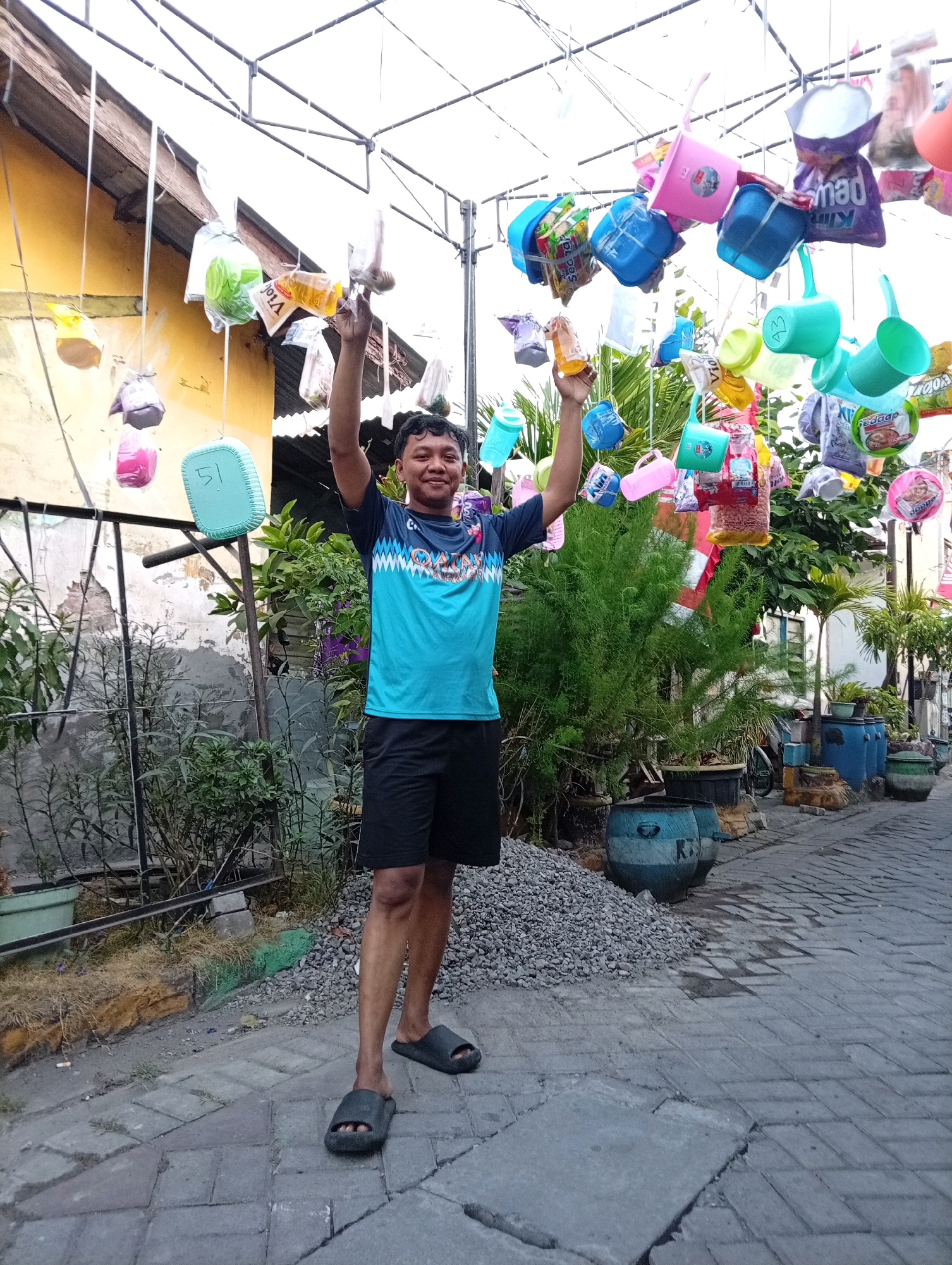 In Surabaya, Indonesia, a man stands outdoors with his arms raised. He is wearing a blue and teal t-shirt with a patterned design and black shorts. The man is smiling and looking at the camera. He is standing on a paved walkway, and in front of him, a pile of grey gravel. Behind him, a building with a yellow facade and a metal fence. Hanging above the man are numerous colorful plastic containers, bags of snacks, and bottles, suspended by wires, creating an artistic display. Lush green plants and large blue planters are also visible in the background, suggesting a decorative or community space.