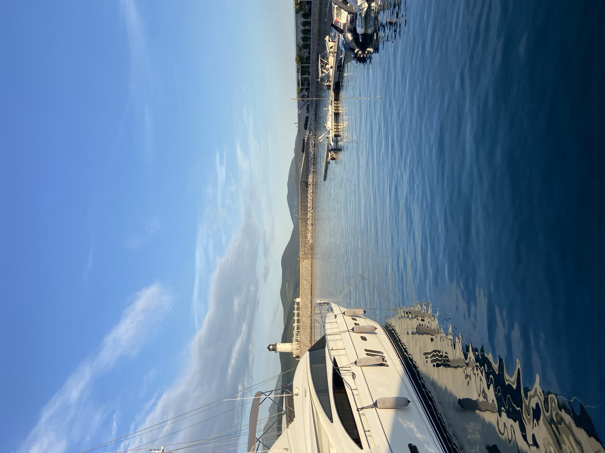 The image captures a serene marina scene on a bright, sunny day in Radovići, Montenegro. A large, elegant white motor yacht, partially visible in the foreground on the right, dominates the composition. Its sleek design features dark windows and several white fenders hanging along its side, reflecting in the calm, deep blue water.
Extending into the water from the center, a long stone breakwater or pier is visible, serving as a docking area for numerous other boats and yachts further in the distance. A white lighthouse stands prominently on this pier, behind the foreground yacht. Beyond the pier, arched structures, possibly part of a waterfront building or aqueduct, are discernible on the land. In the background, a range of gentle mountains or hills stretches across the horizon, complementing the coastal landscape.
The sky is a vibrant blue, adorned with scattered, wispy white clouds, suggesting clear and pleasant weather. The water is notably still, reflecting the sky and creating rippled, abstract patterns of the boats and structures along the pier. The strong, clear lighting indicates a late morning or early afternoon setting. No people are prominently visible, contributing to the tranquil atmosphere.
Visible text on the side of the main yacht reads "AXES 63". On a smaller vessel docked further away, the word "TARGA" is identifiable. The overall scene evokes a sense of peace and leisure in a picturesque Mediterranean setting.