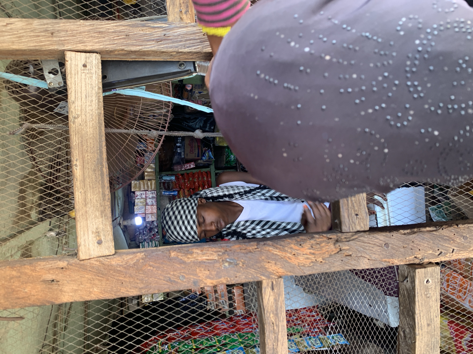 A man with a black and white checkered headscarf and a white shirt is sleeping on a shelf inside a small shop. He is viewed from above through a wooden and wire mesh structure. Various items are visible on the shelves of the shop, including snacks and drinks. A fan and a lightbulb are also present. Part of a person's body, wearing a patterned gray garment, is in the foreground, partially obscuring the view. The location appears to be a small retail establishment, possibly in Lokoja, Nigeria.