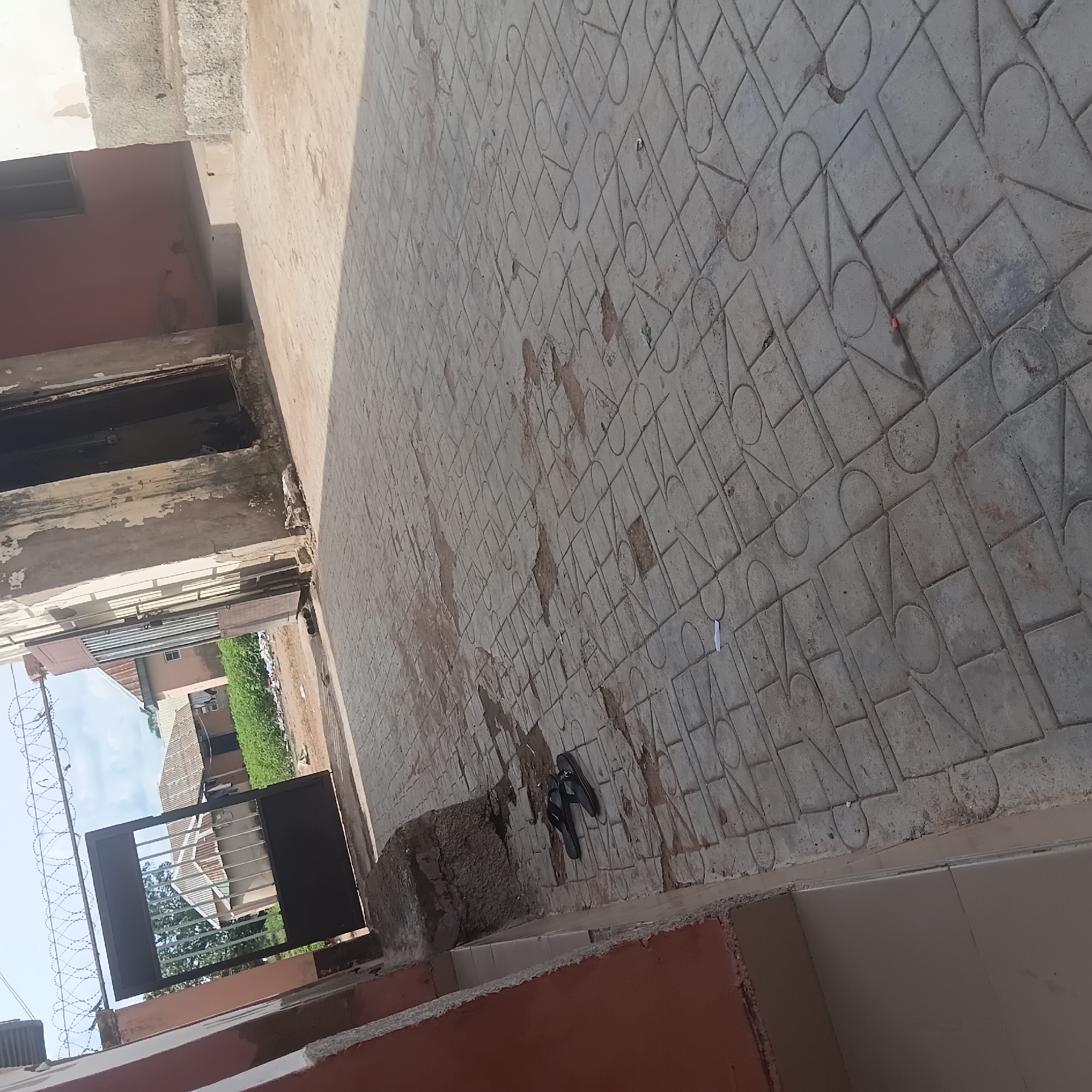 The image depicts an outdoor scene in Wukari, Nigeria, featuring a paved walkway in front of a building with a gate. The walkway is constructed of patterned concrete interlocking pavers. To the left, a building wall painted in a reddish-brown hue is visible. A large, dark metal gate, partially open, is situated at the entrance, topped with barbed wire. Beyond the gate, residential buildings with corrugated metal roofs and surrounding vegetation can be seen. In the foreground, on the paved walkway, a pair of black flip-flops are placed. The ground shows some areas of wear and tear on the pavers, with some dirt and debris scattered about. The overall impression is of a somewhat aged urban or semi-urban environment.