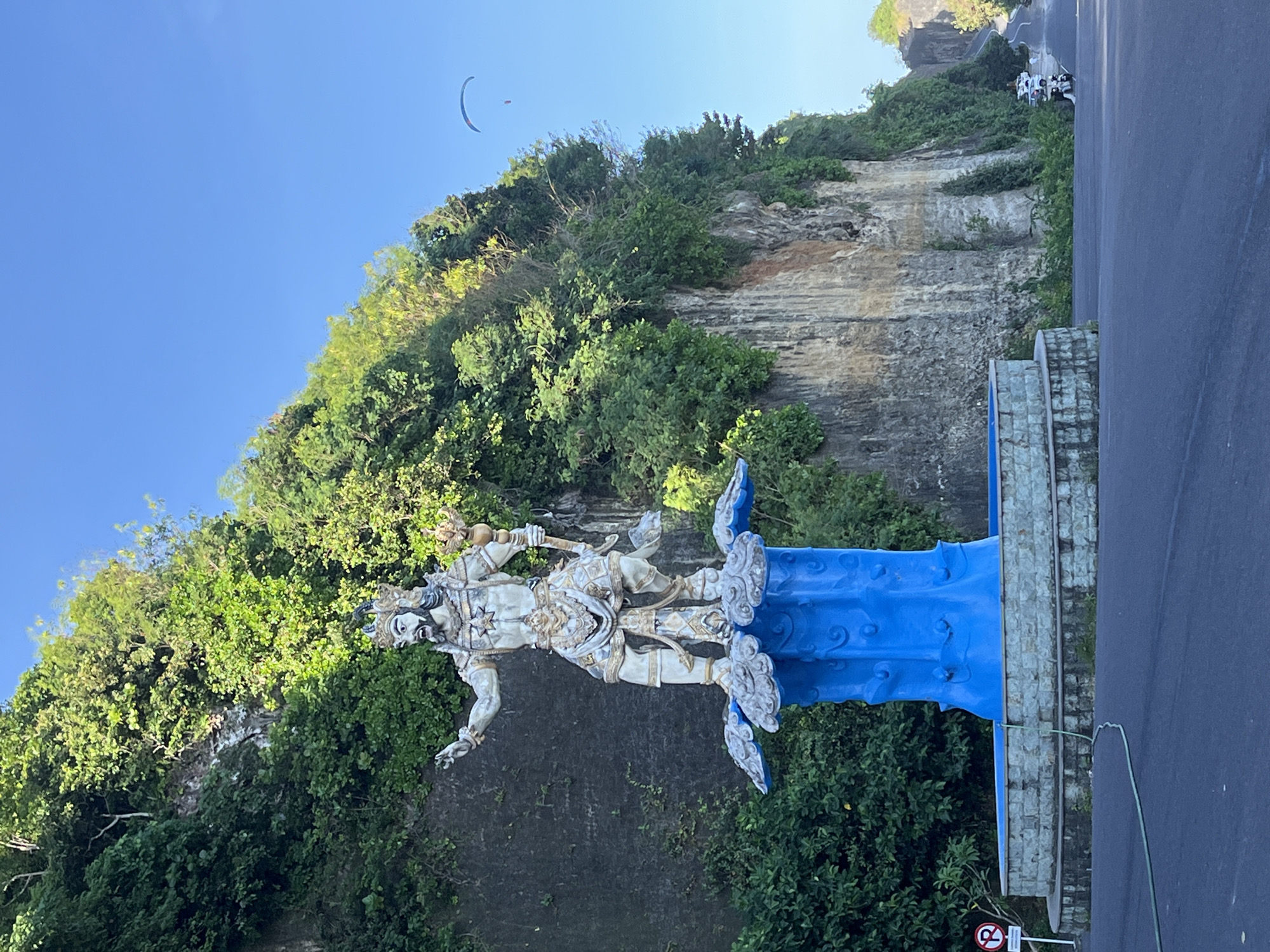 The image shows a statue of an Indonesian deity in Kuta Selatan, Indonesia. The statue is white and stands on a blue pedestal adorned with cloud-like designs. The statue is positioned in front of a cliff covered in lush green vegetation. A road runs alongside the statue. In the upper part of the image, a paraglider is visible in the blue sky. Several parked motorcycles are visible further up on the road.