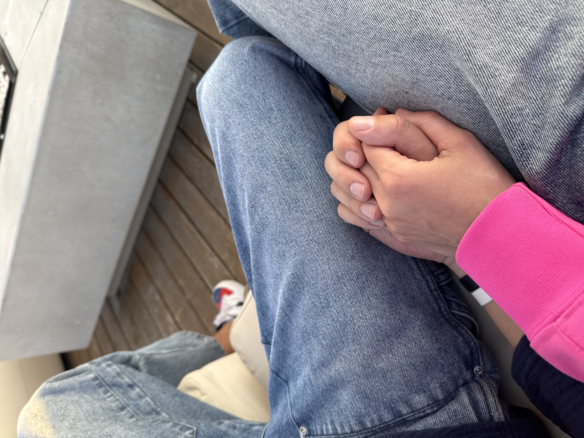 The image shows a close-up, overhead view of a person seated with their hands clasped on their lap. The person is wearing blue jeans, and their hands are resting on their legs. Part of another person is visible, wearing jeans and a bright pink long-sleeved shirt. A white and red shoe is visible in the lower left corner. The setting appears to be outdoors, possibly on a wooden deck, next to a concrete structure. There is no information that indicates the location is in Мысовское сельское поселение, Ukraine.