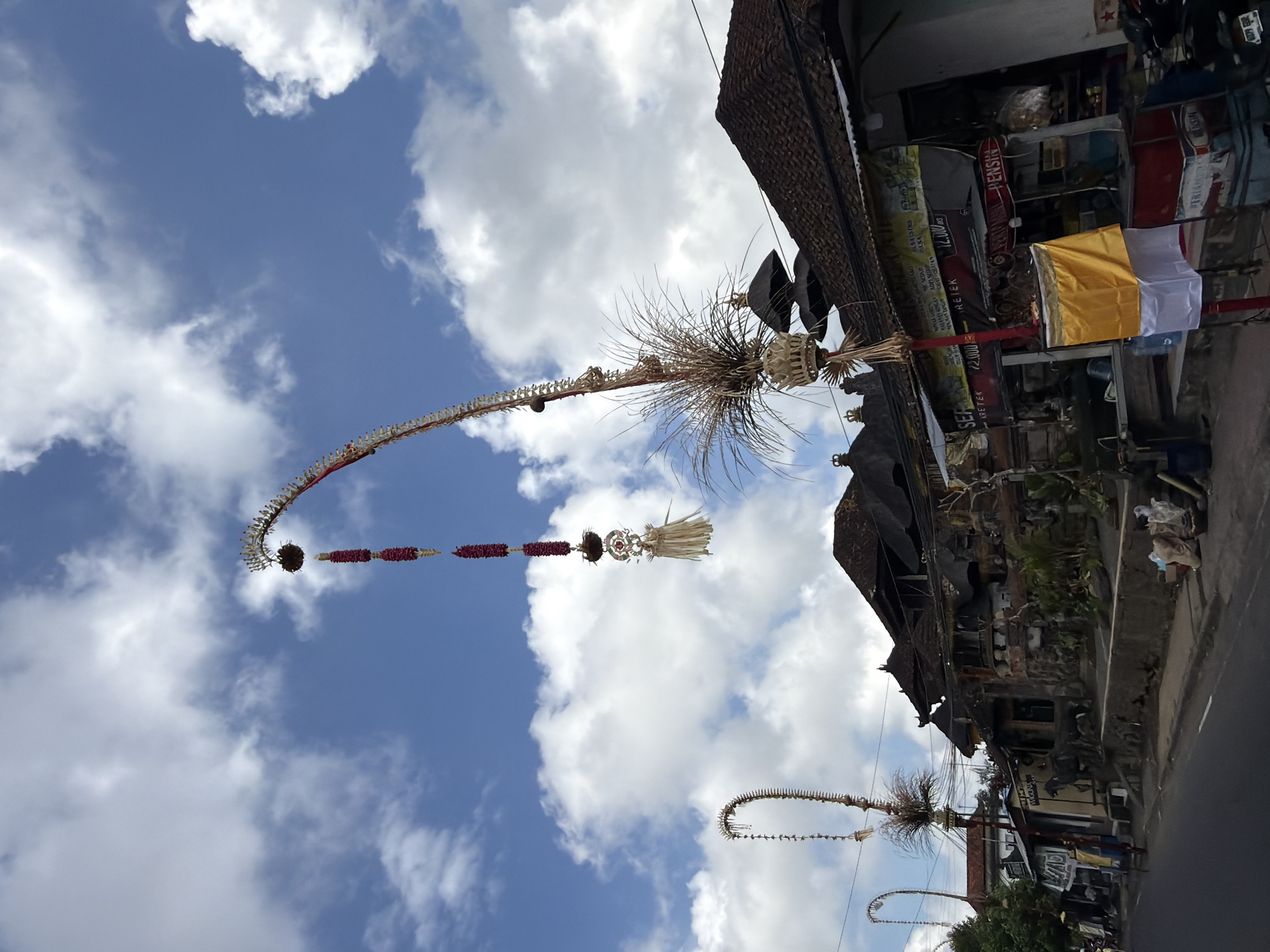 In Tampaksiring, Indonesia, a low-angle, full shot captures a street scene under a partly cloudy blue sky. Two elaborately decorated tall, curved poles, known as Penjor, stand prominently in the foreground, adorned with red flower garlands and natural materials like straw and dried leaves. One Penjor is positioned to the left, its curved form reaching towards the sky, while another, larger Penjor dominates the center-right of the frame, its intricate top section featuring woven elements and a feathery, grass-like decoration.
Beneath the Penjor, traditional Balinese architecture is visible, characterized by dark, tiled, multi-tiered roofs. The buildings appear to be shops or residences, with signs in Indonesian text, some displaying prices in Rupiah (RS). Evidence of commerce is present, with items like bottles, containers, and bundled goods displayed outside. A few individuals are visible on the street, including a person walking on the left and what appears to be a cart. The road in the foreground is asphalt. The overall impression is one of a vibrant, culturally rich street in an Indonesian town.