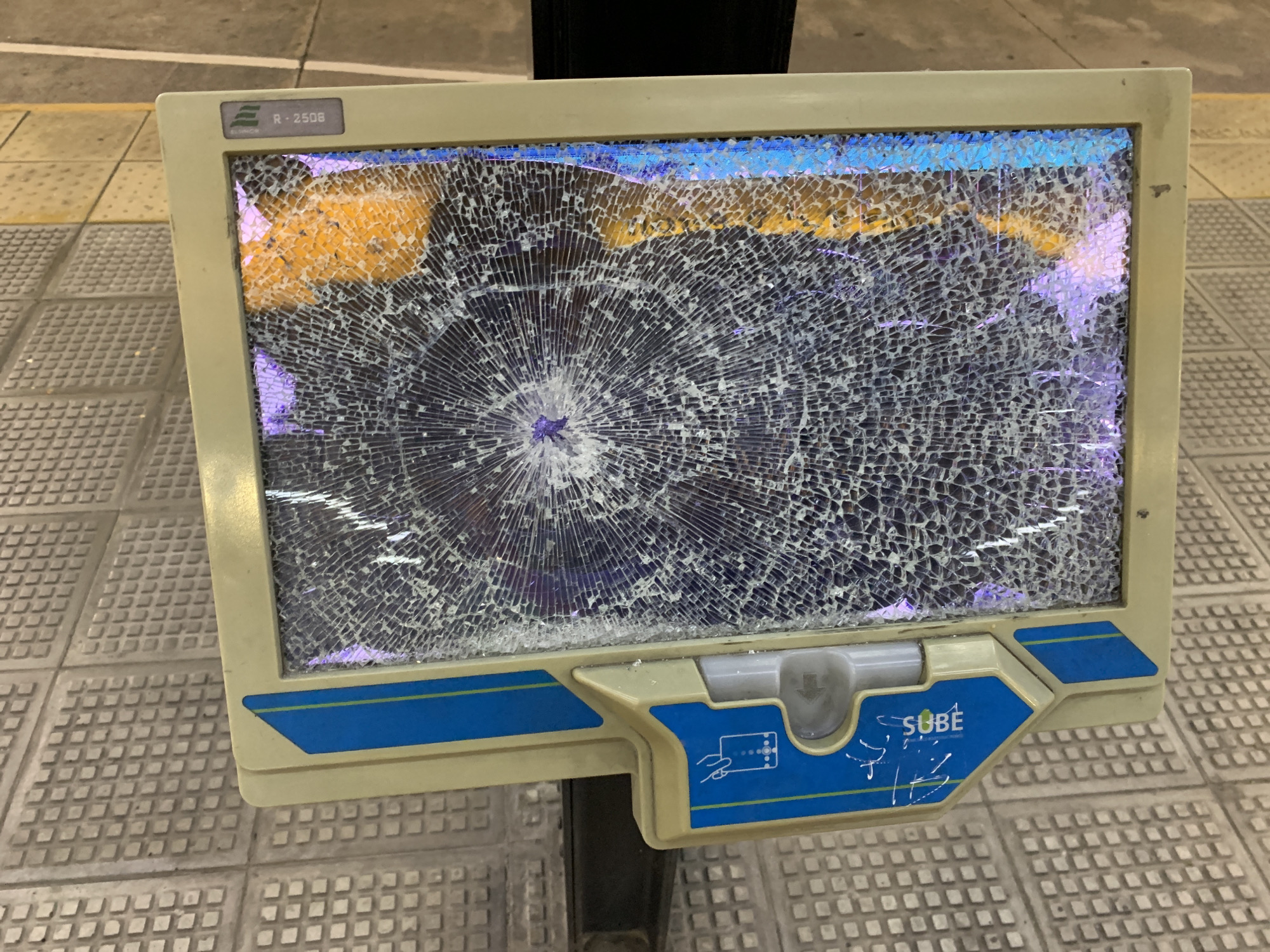 The image shows a damaged SUBE card reader in Buenos Aires, Argentina. The screen is shattered with a spiderweb-like pattern emanating from a central point. The card reader is light beige with blue accents and features the "SUBE" logo. It is mounted on a black pole and stands on a tiled platform. The screen displays an unclear image with traces of yellow and blue color. There's an "R-2508" identifier on the top left. The reader's card slot is visible below the damaged screen. There is some graffiti under the logo.