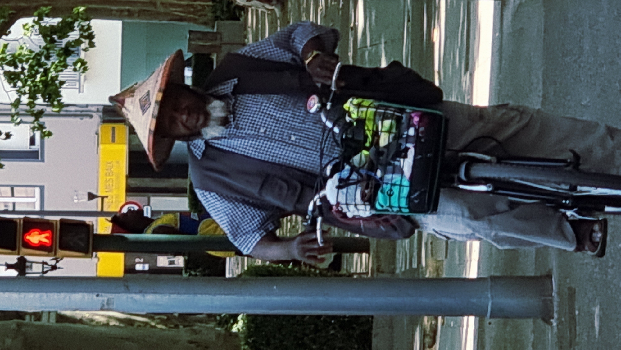 The media file shows a person on a bicycle in what appears to be a street scene in Olot, Spain. The person is wearing a conical hat, a checkered shirt, and a dark vest. They are holding onto the handlebars of the bicycle, which has a basket attached to it filled with various items, including what appear to be plastic bottles and fabric. The person's right hand is also visible, and they appear to be holding a cigarette. The bicycle has a rear rack and a dark-colored frame.
In the background, there is a traffic light showing a red pedestrian signal, indicating it is not safe to cross. There are also buildings visible, one of which has a yellow sign with text that is partially legible. The overall scene suggests a daytime setting, likely outdoors, with the sun casting shadows, hinting at a sunny day. The presence of a traffic light and buildings suggests an urban or semi-urban environment. The person's expression is not clearly discernible due to the angle and quality of the image, but their posture suggests they are in motion or stopped momentarily. There is also a grey pole in the foreground, possibly a lamppost or part of street furniture.
