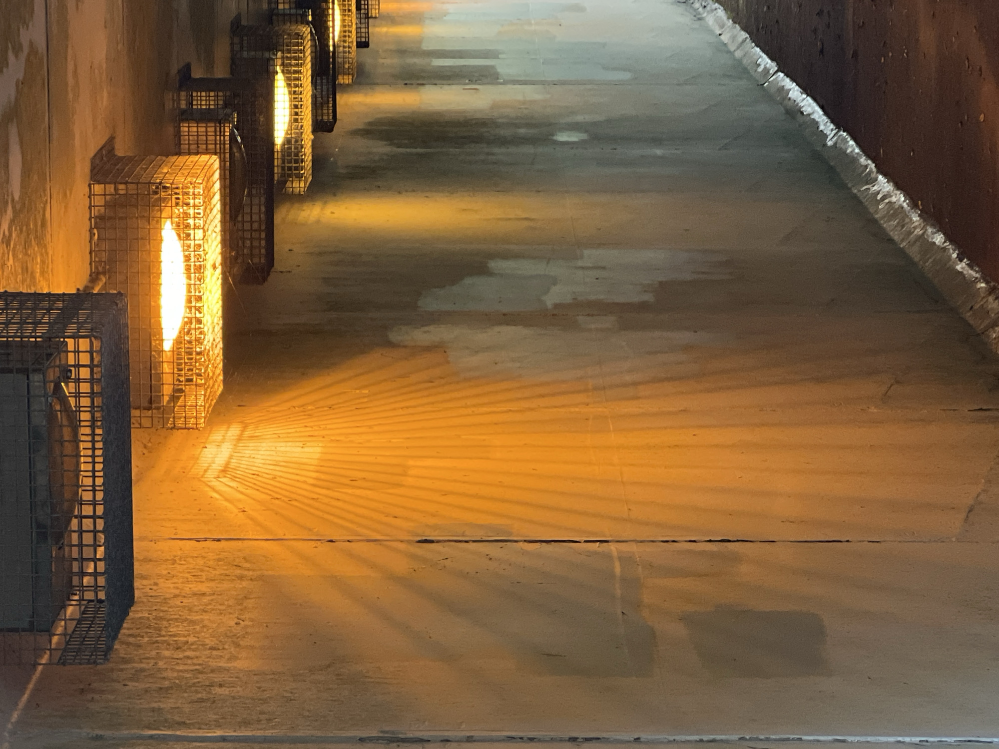 The image depicts a concrete wall illuminated by multiple lights, creating dramatic shadows. The lights are housed in wire cages and are mounted on the ceiling, which is also concrete. The wall has a mottled appearance with streaks and discolored patches, suggesting it is exposed to the elements. The lighting casts a warm, yellowish glow, with one prominent beam creating a fan-like pattern of shadows on the wall. The ground visible in the lower right corner is a dark, rough surface, possibly dirt or gravel. The scene appears to be an underpass or a similar infrastructural space within the city of Mountcharles, Ireland. There are no people or animals visible in the image.