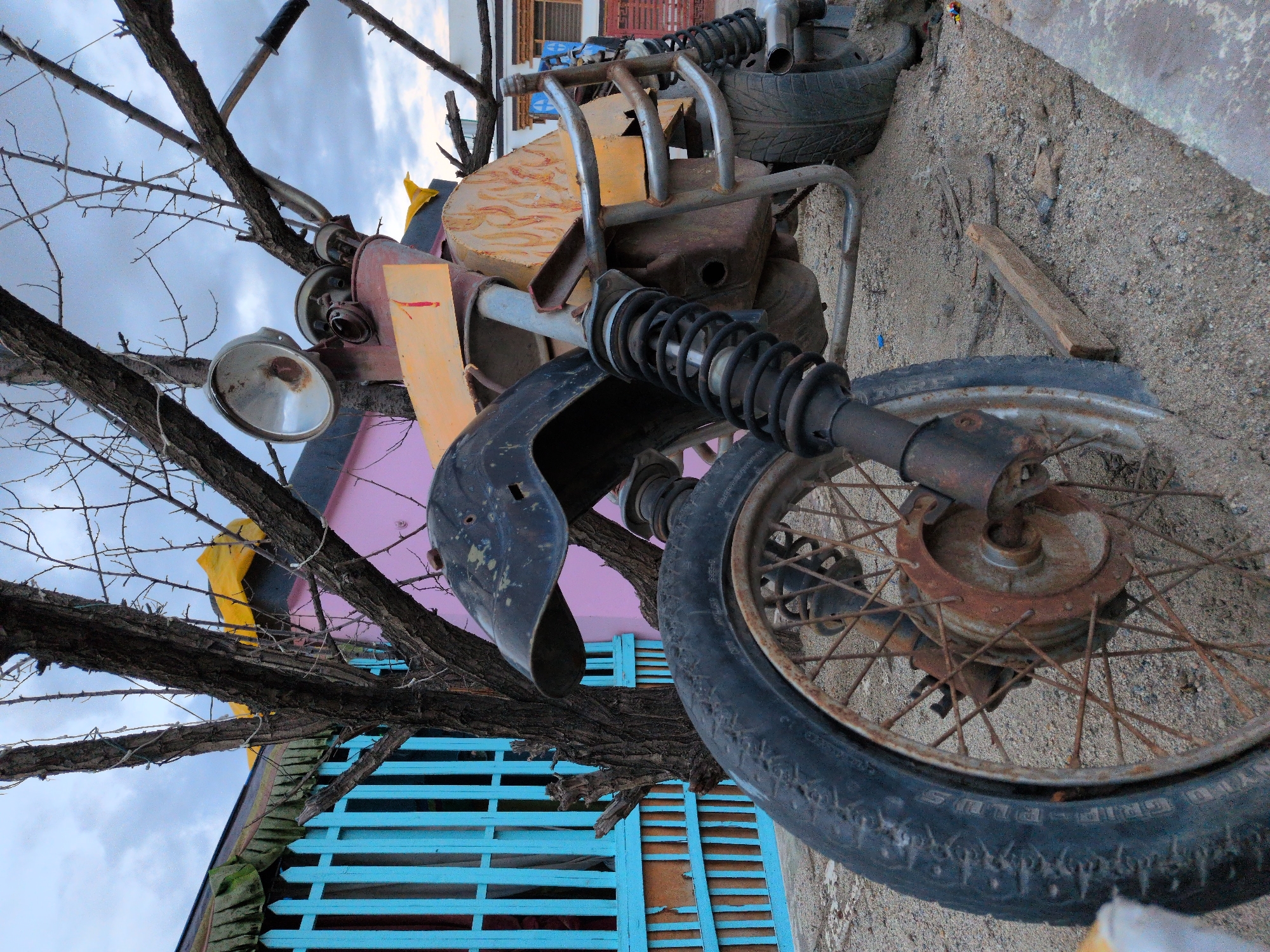 An exterior, eye-level, medium shot captures a heavily rusted and modified motorcycle, partially buried in sand, with a bare tree in the foreground. The motorcycle appears to be a custom build, with various metal components welded together, including a yellow fuel tank with painted flames, and a black fender. The front wheel is clearly visible, with a spoked rim and a worn tire. The background features a bright blue wooden fence and a pink building, suggesting an urban or semi-urban environment. The sky is overcast with clouds. The scene is located in the city of Leh, India. No people are present.