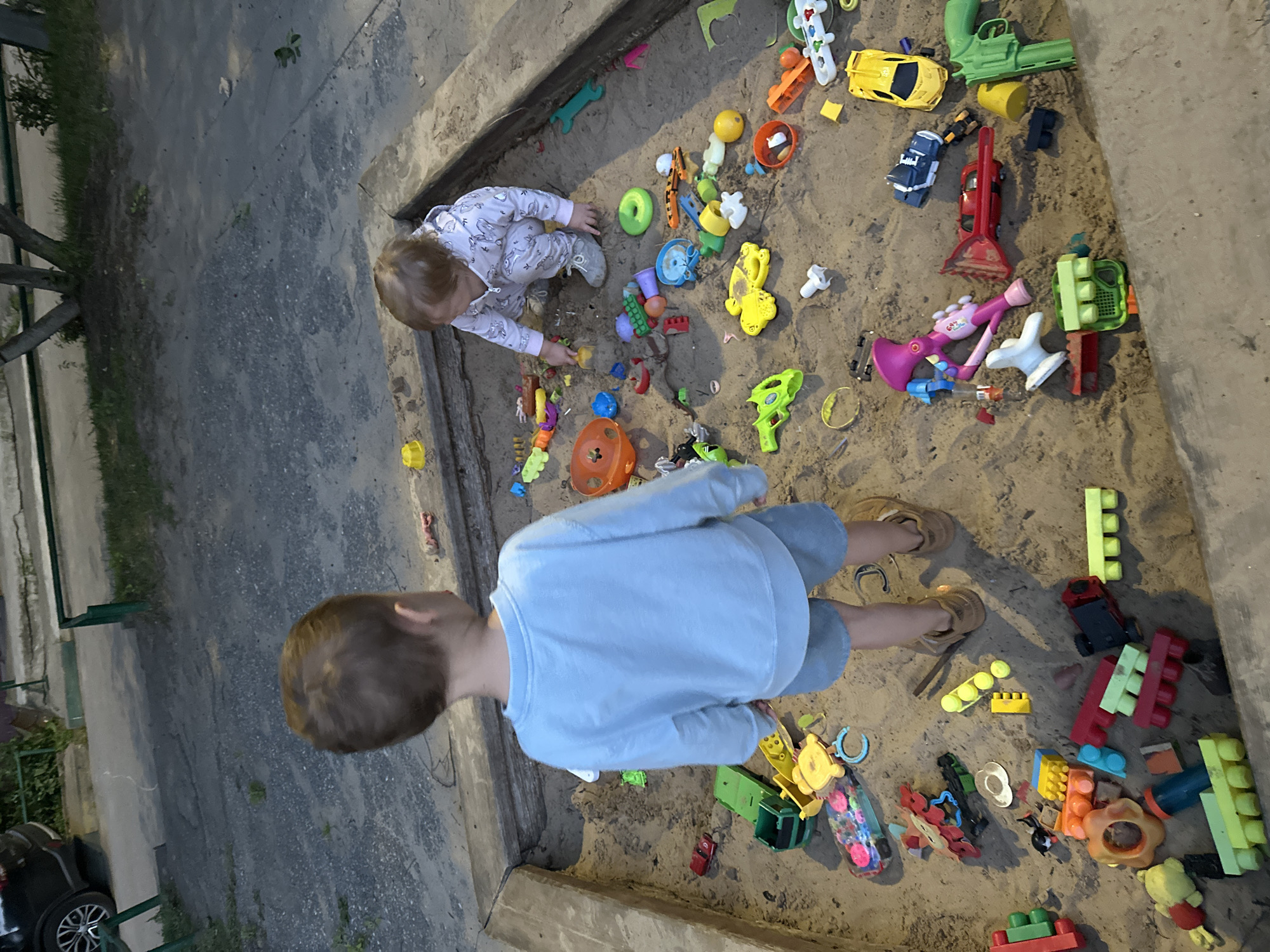 Two children are playing in a sandbox filled with toys. The sandbox is made of concrete and wood. The ground surrounding the sandbox is paved with gravel and concrete. In the background, there is a car parked and some vegetation. The scene is likely outdoors in a residential area.