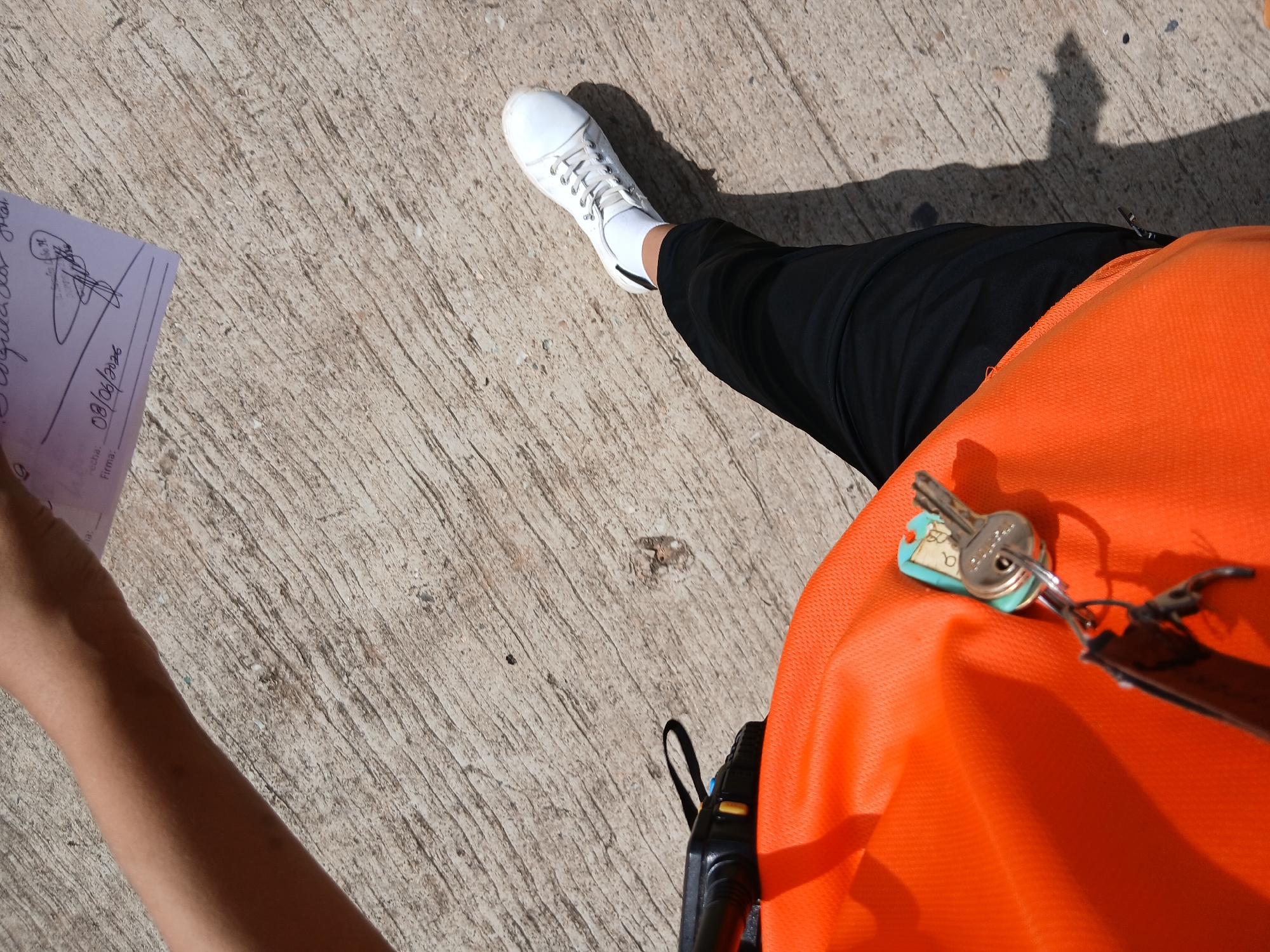 The image, taken at Playa El Agua, Venezuela, shows a first-person perspective of a person standing on a concrete surface. The person is wearing black pants and a white athletic shoe with white socks. An orange safety vest or shirt is visible, with a small radio clipped to it. A set of keys hangs from a turquoise tag attached to the vest. In the upper portion of the image, a hand is holding a piece of paper with handwriting and a signature, dated "08/06/2026". The concrete surface appears weathered and textured, typical of an outdoor area.