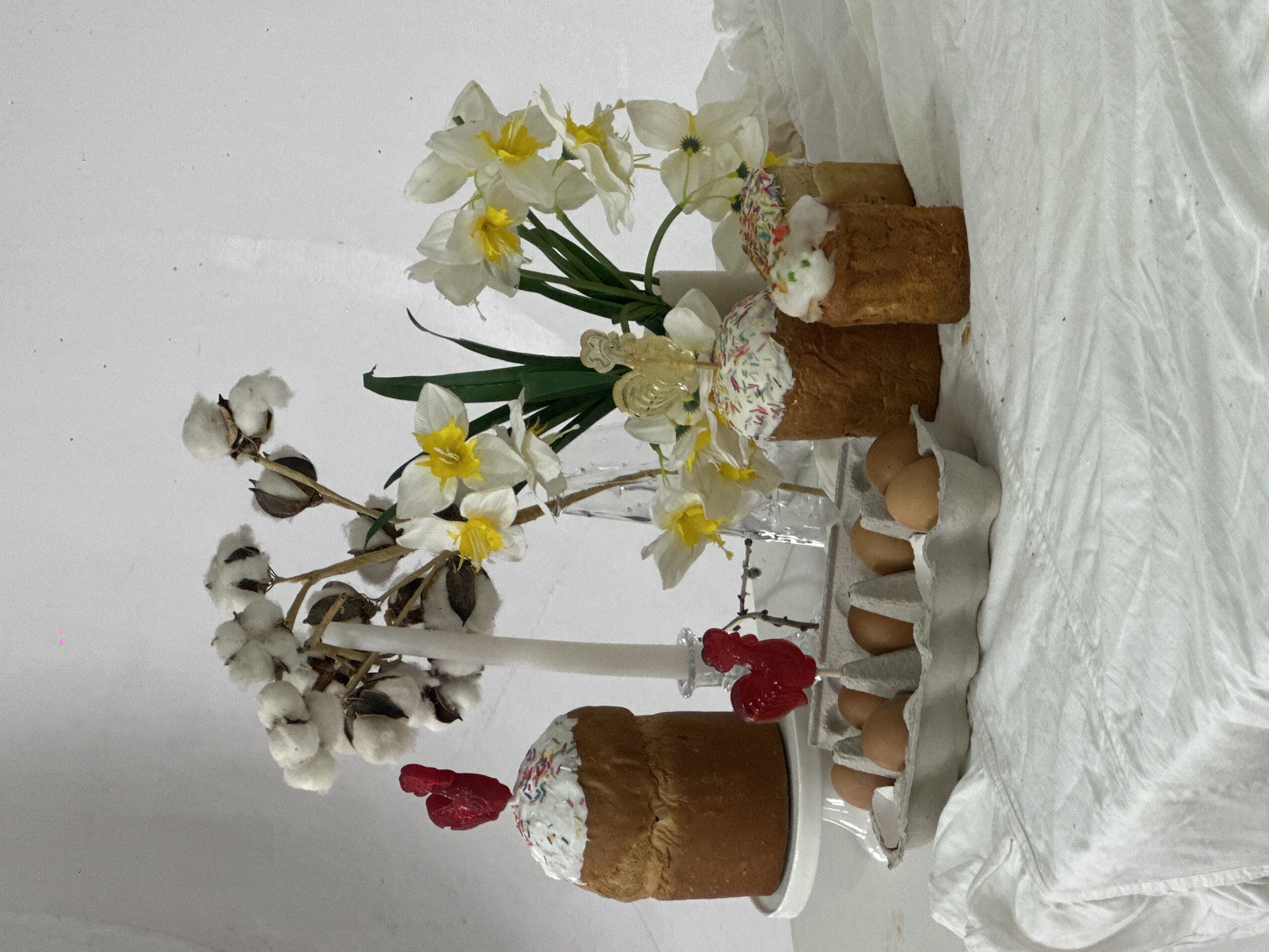 The image presents an Easter still life, likely set indoors. Three Easter cakes, adorned with white icing and colorful sprinkles, are arranged among other items. A sprig of cotton flowers, daffodil blossoms, a white candle with red ornaments, and a carton of brown eggs are part of the composition. The scene is set against a white backdrop, part of which is draped with white fabric. The location is likely the city of Chaikovsky, Russia.