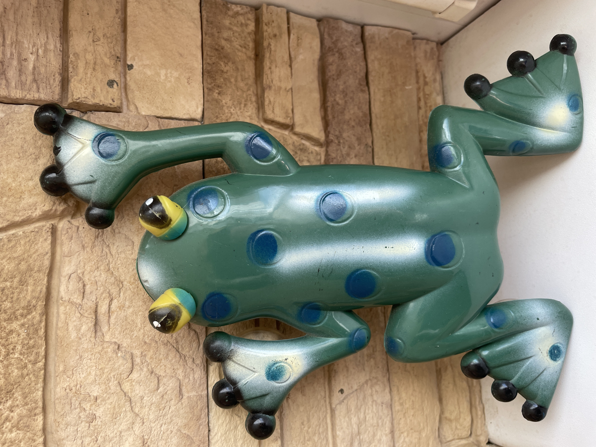 A green, plastic frog decoration is mounted on a textured, brick wall. The frog has blue spots, yellow eyes, and black toes. The location is unknown.